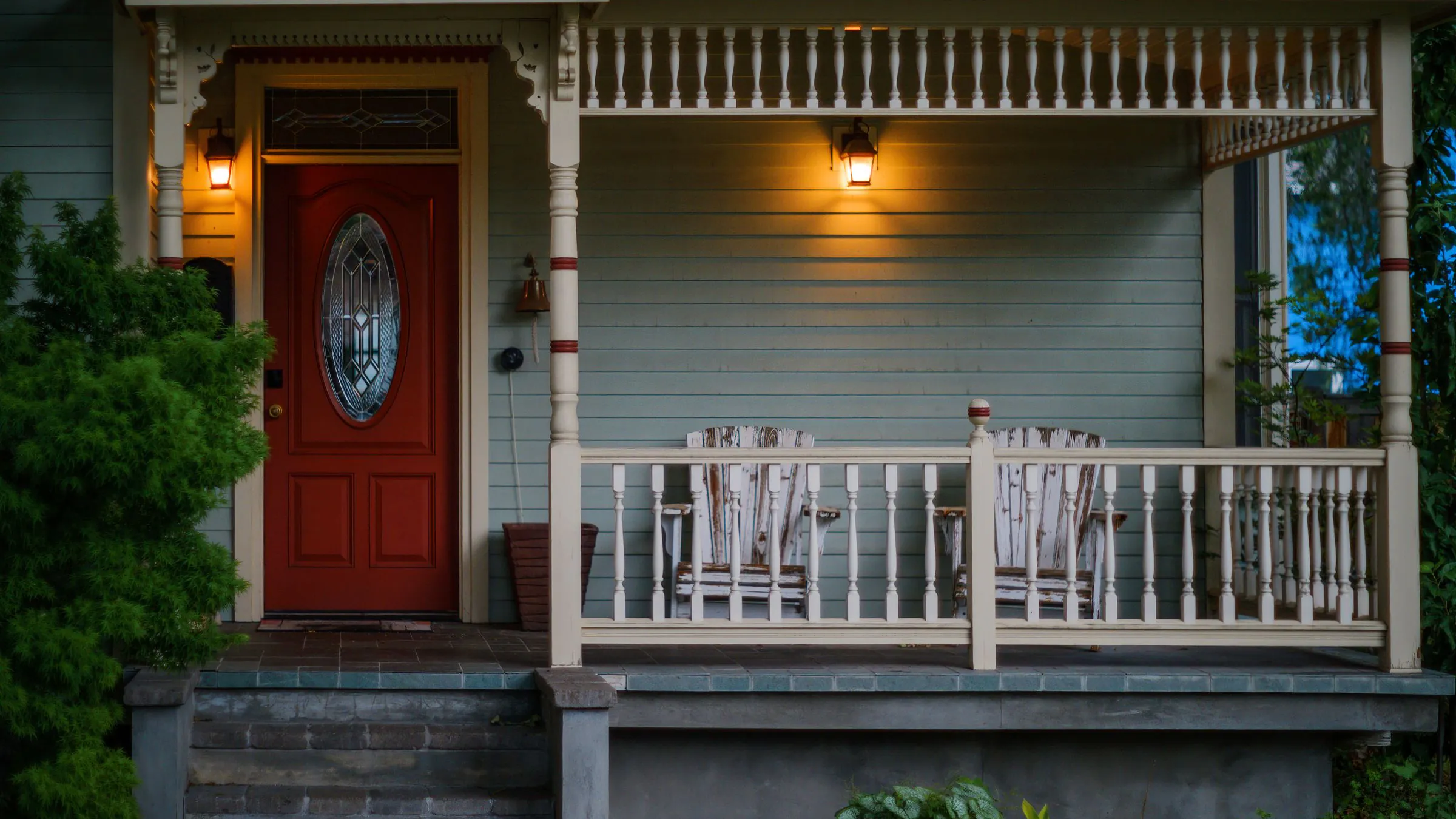A red front door and a covered porch with two wooden chairs, lit by wall lamps at dusk