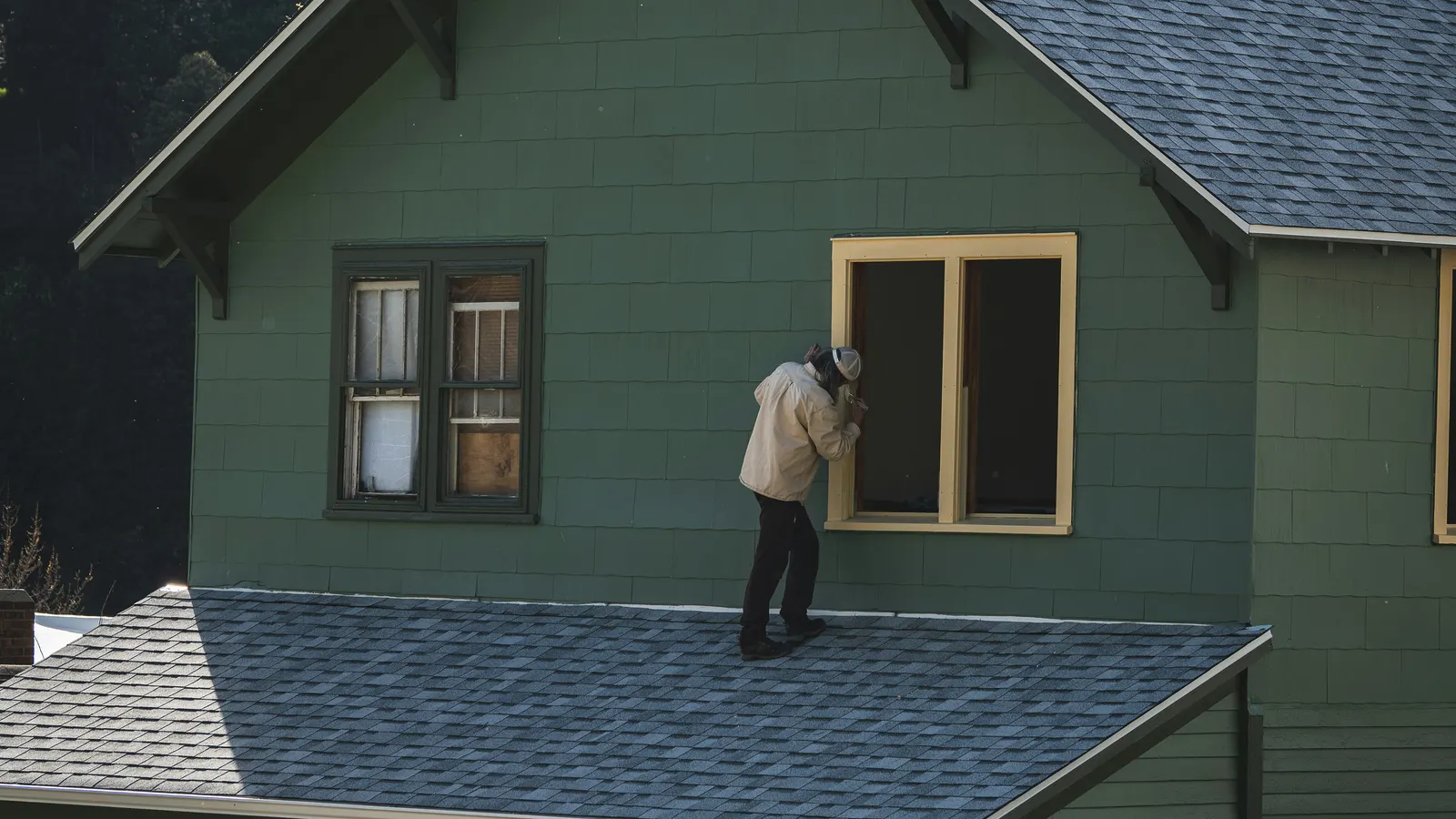 A painter seen from behind stands on an asphalt-shingle porch roof, brushing paint onto the cream trim of an upstairs window on a green shingle-sided house in clear daylight
