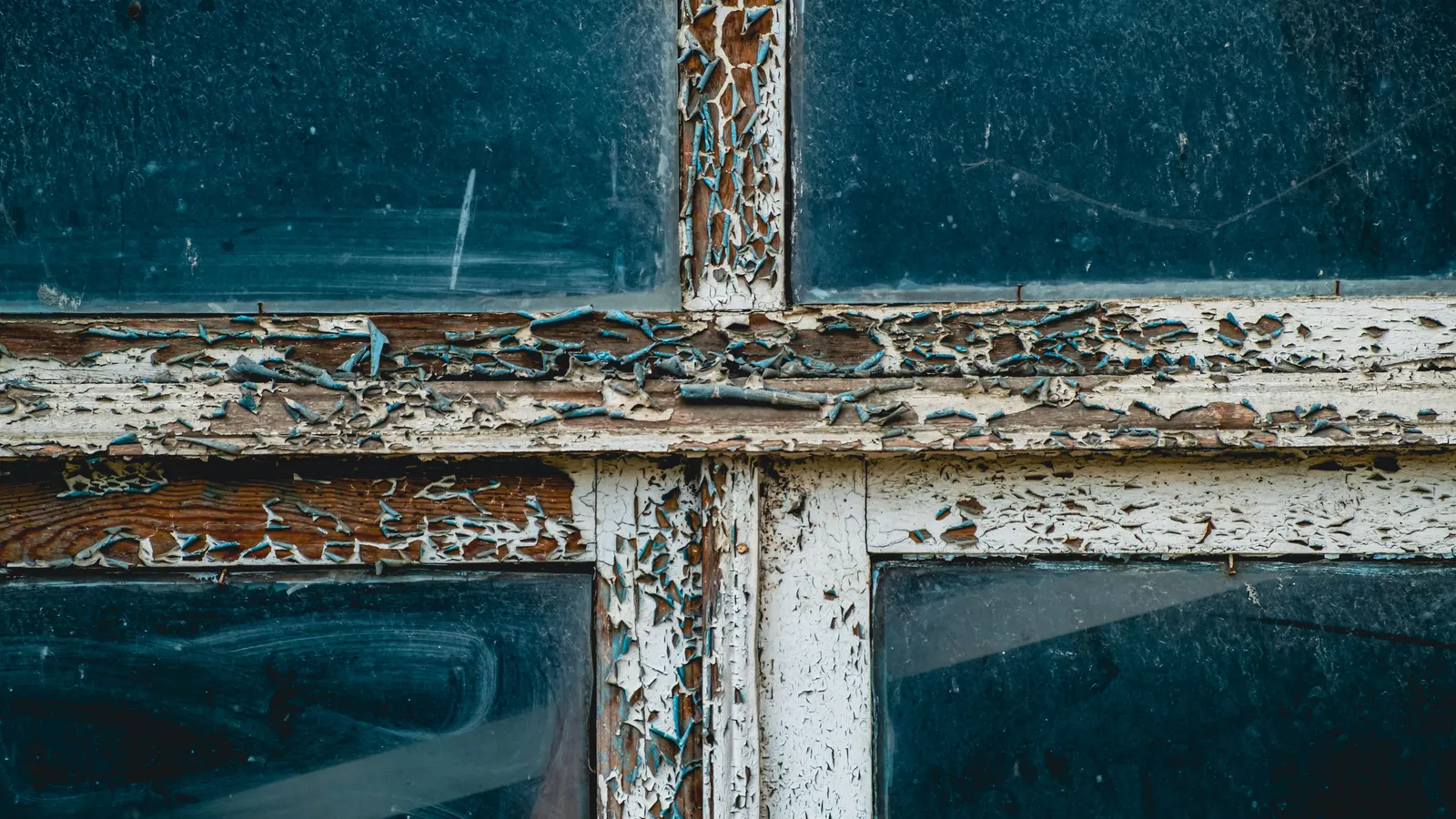 Close-up of an old wooden window frame with white and blue paint cracking and flaking away from the weathered wood underneath