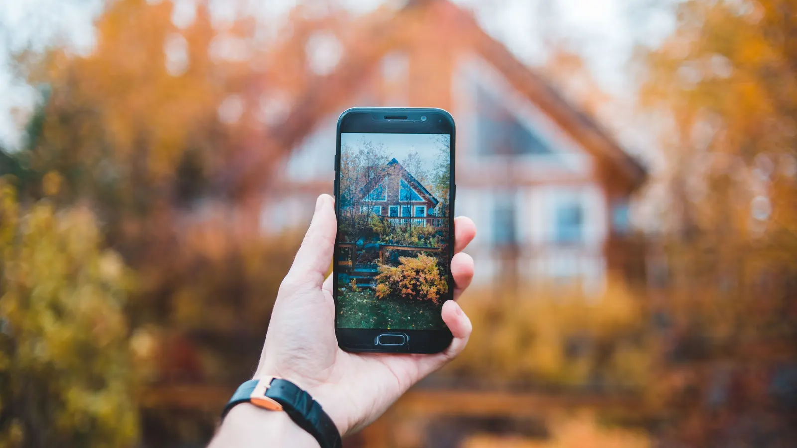 A hand holds up a smartphone showing a photo of a gabled wood-sided house, with the same house blurred behind it among autumn trees