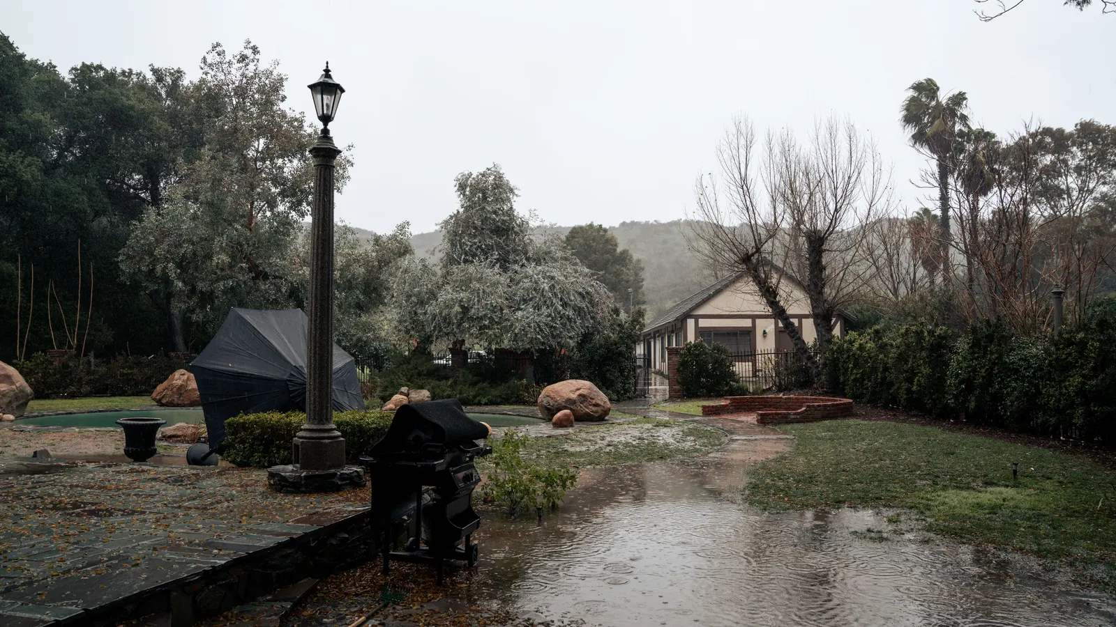 Rainwater pooling across a backyard lawn and stone patio during a storm, with a small house, trees and hills behind