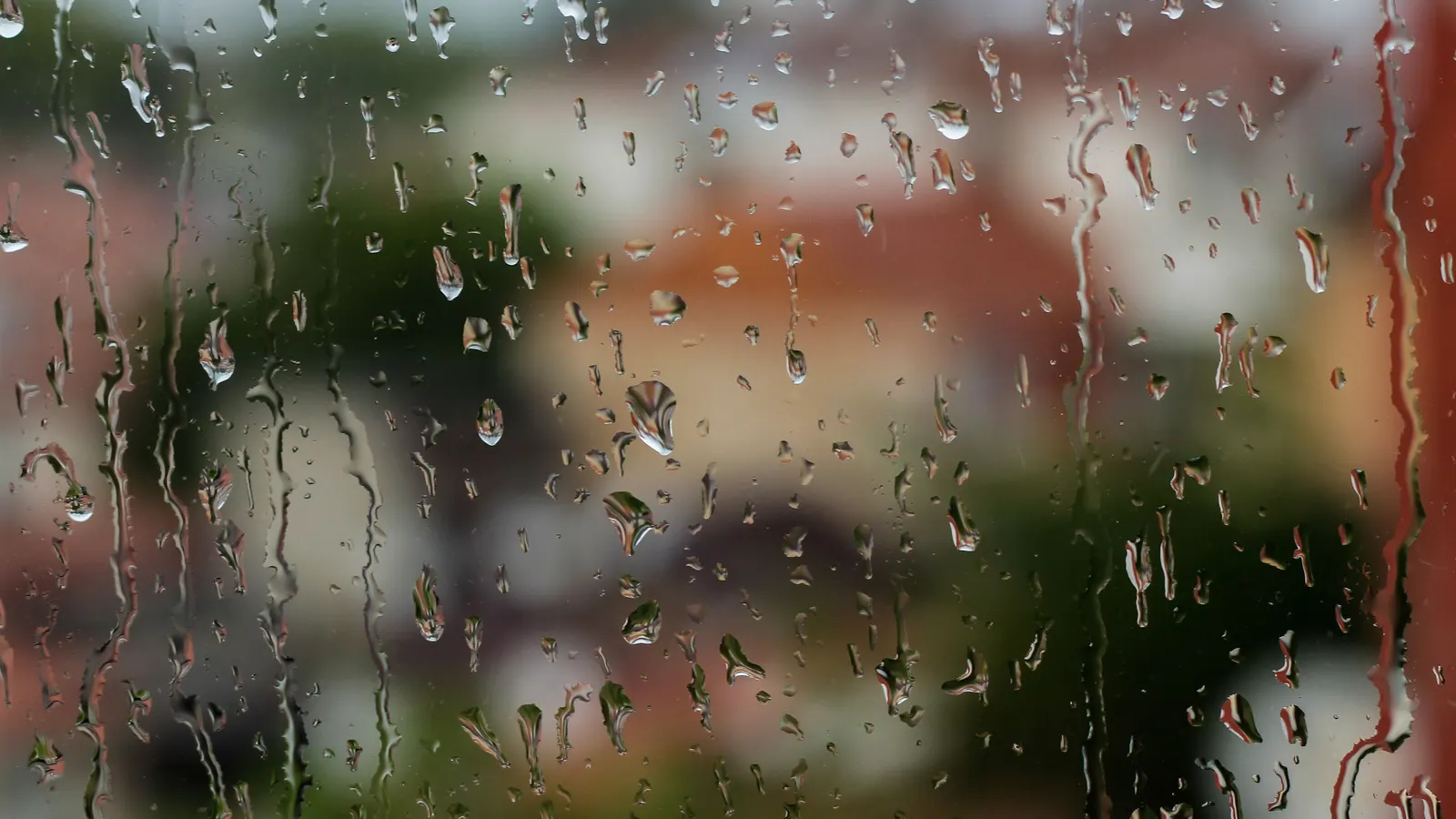 Raindrops streaming down a window pane, with blurred houses and green trees outside