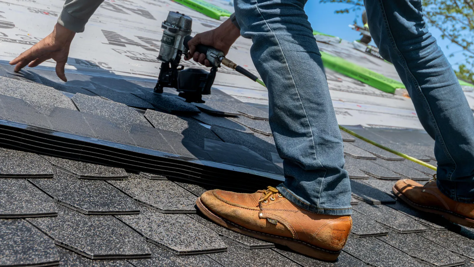 Hands setting a new asphalt shingle in place with a nail gun, work boots braced on the finished rows below