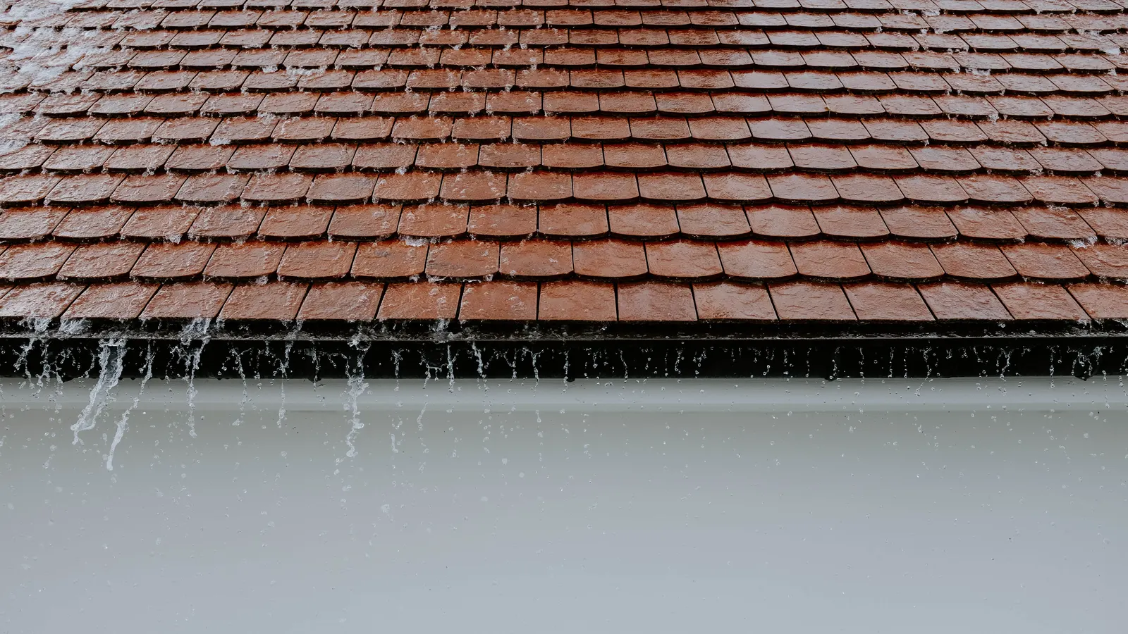 Rainwater streaming over the edge of a wet tiled roof and dripping past the eave in a steady rain