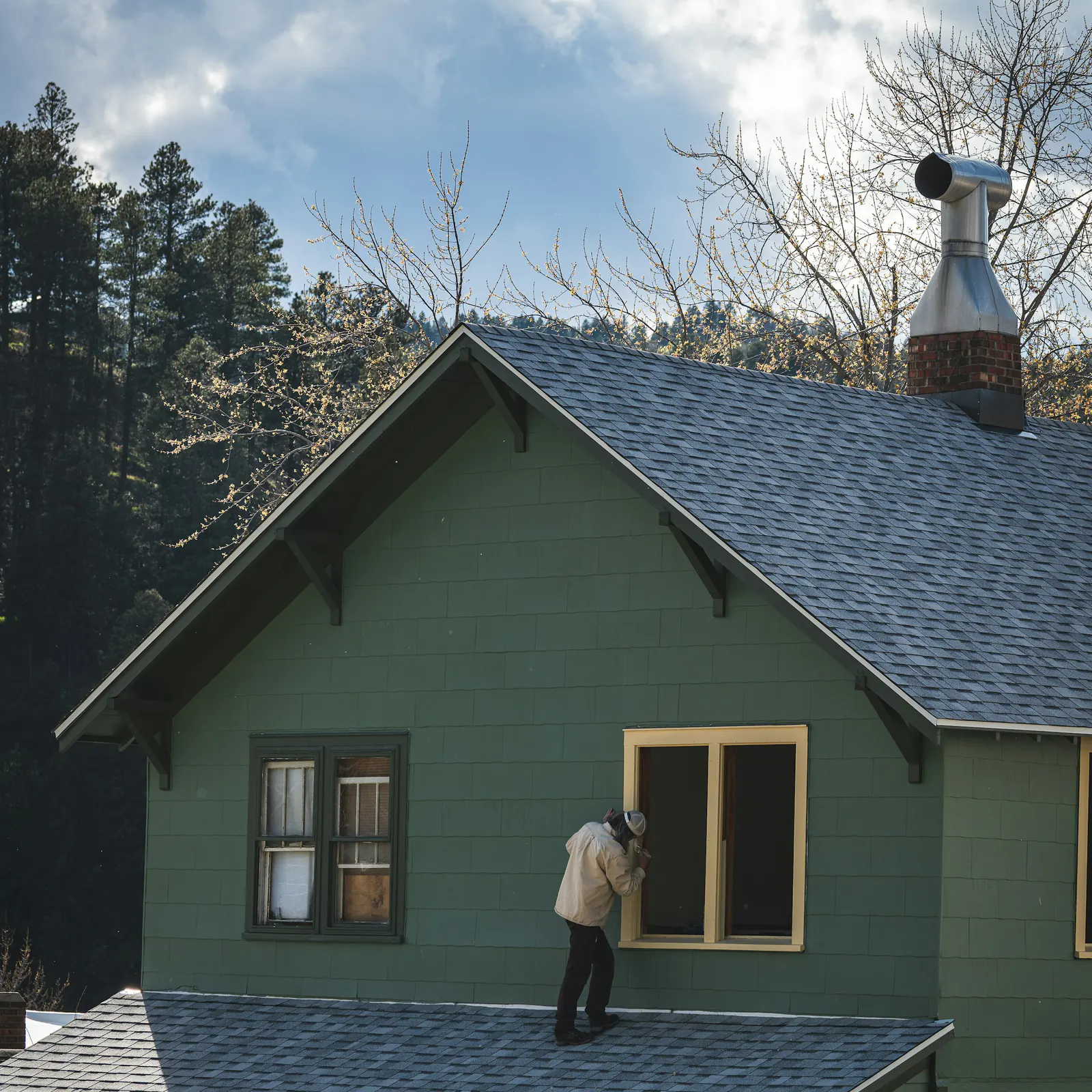 A painter on a porch roof painting the trim of a window on a green shingled Craftsman house