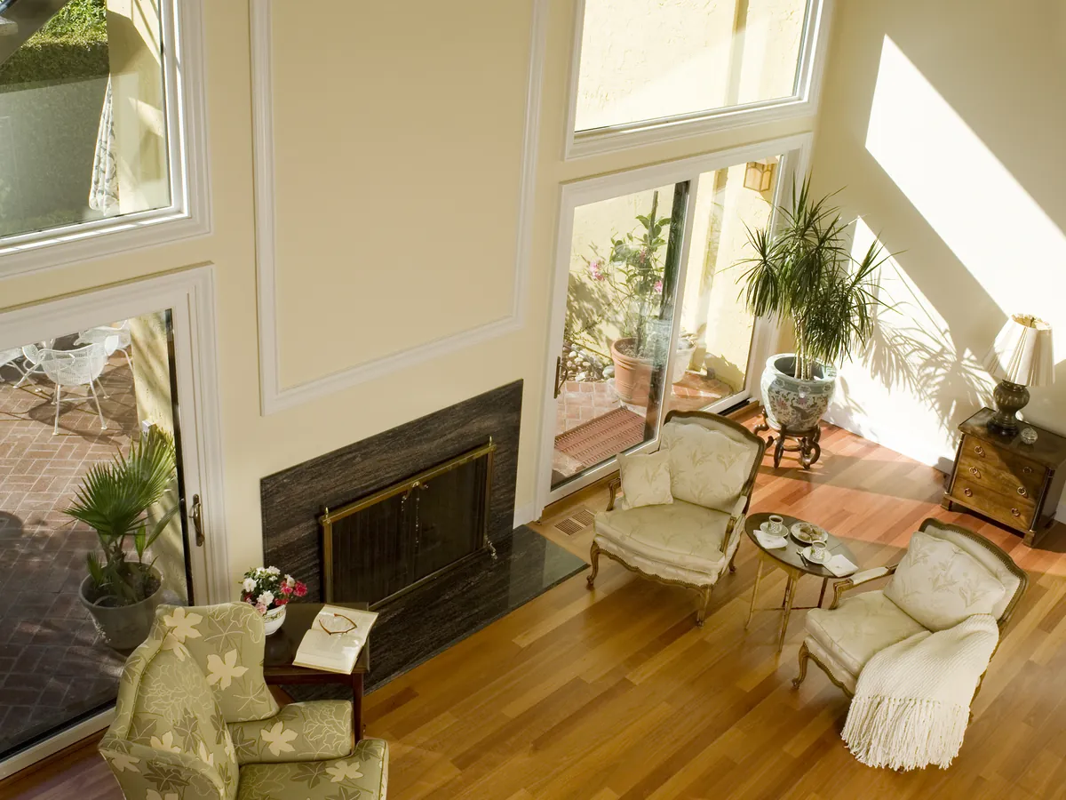 A sunlit two-story living room with warm yellow walls, a black fireplace and pale armchairs