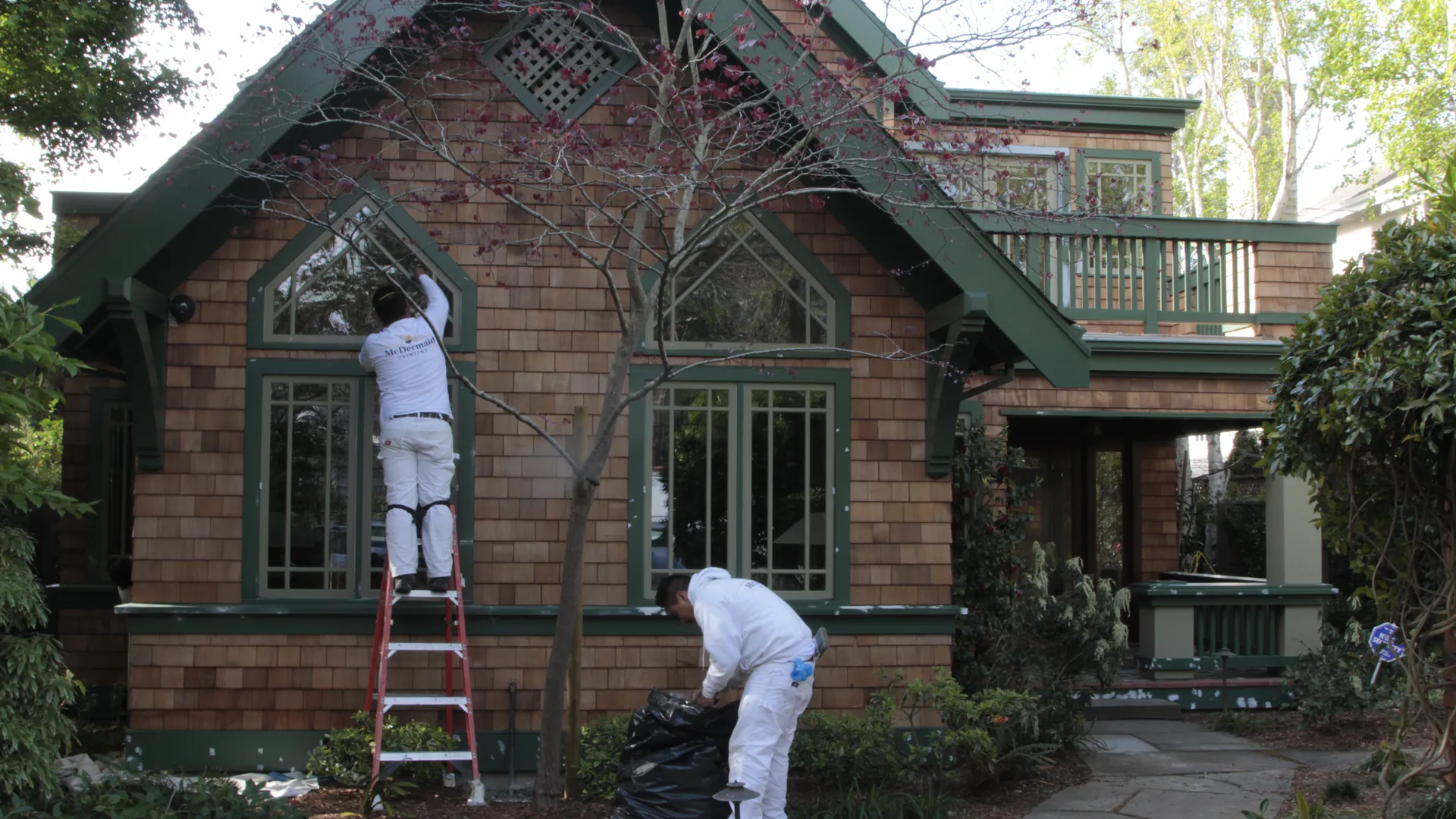 A green-trimmed cedar-shingle gabled cottage with a painter at work on a ladder