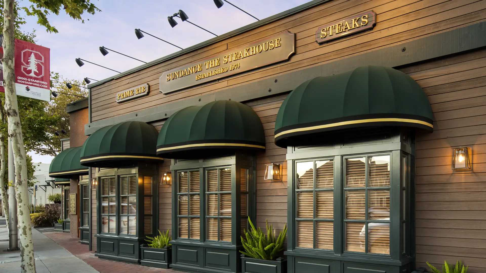 A long restaurant facade with dark green awnings and gold signage in daylight