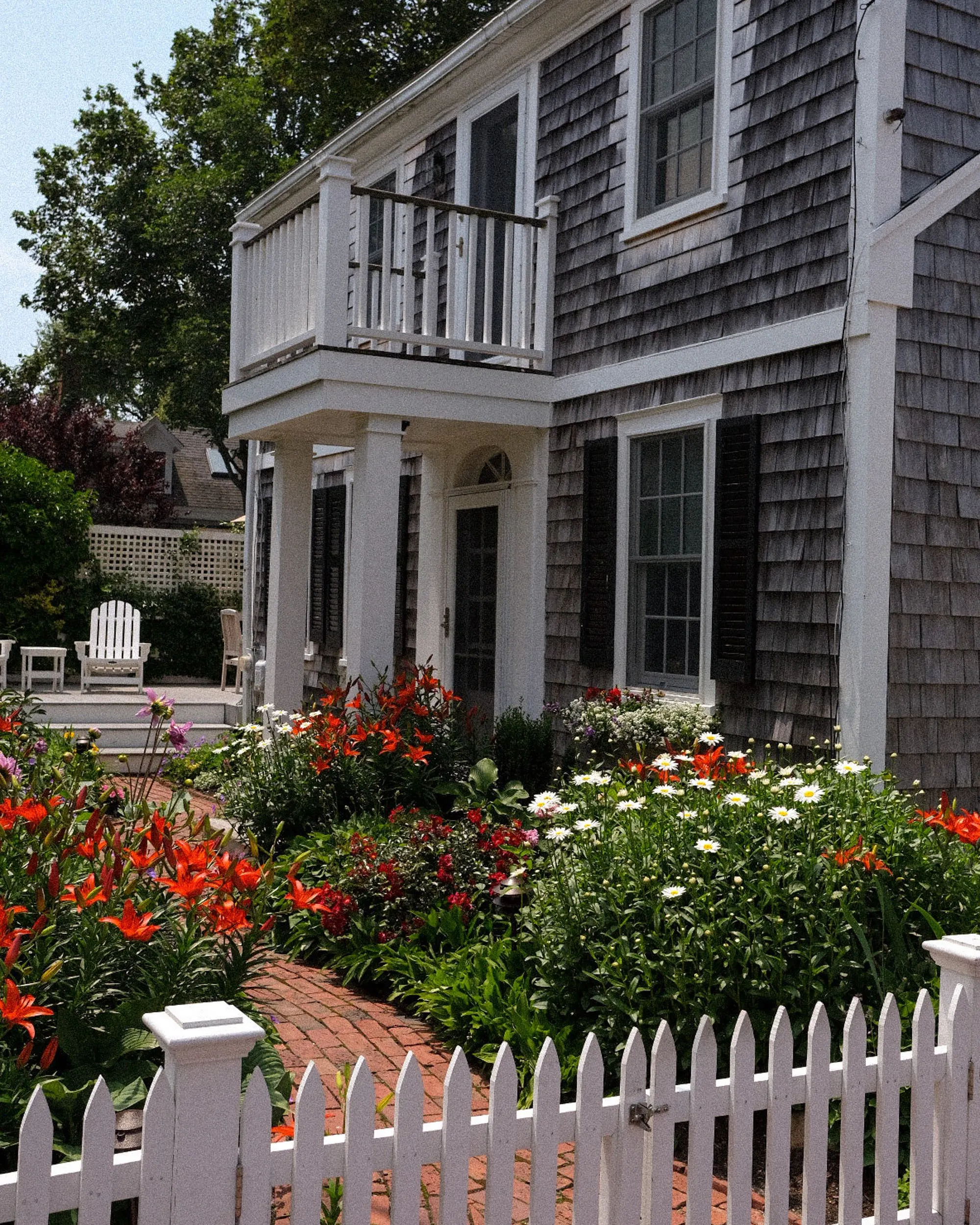 A grey shingled two-storey house with a white balcony and porch, behind a picket fence and a garden of red lilies and white daisies