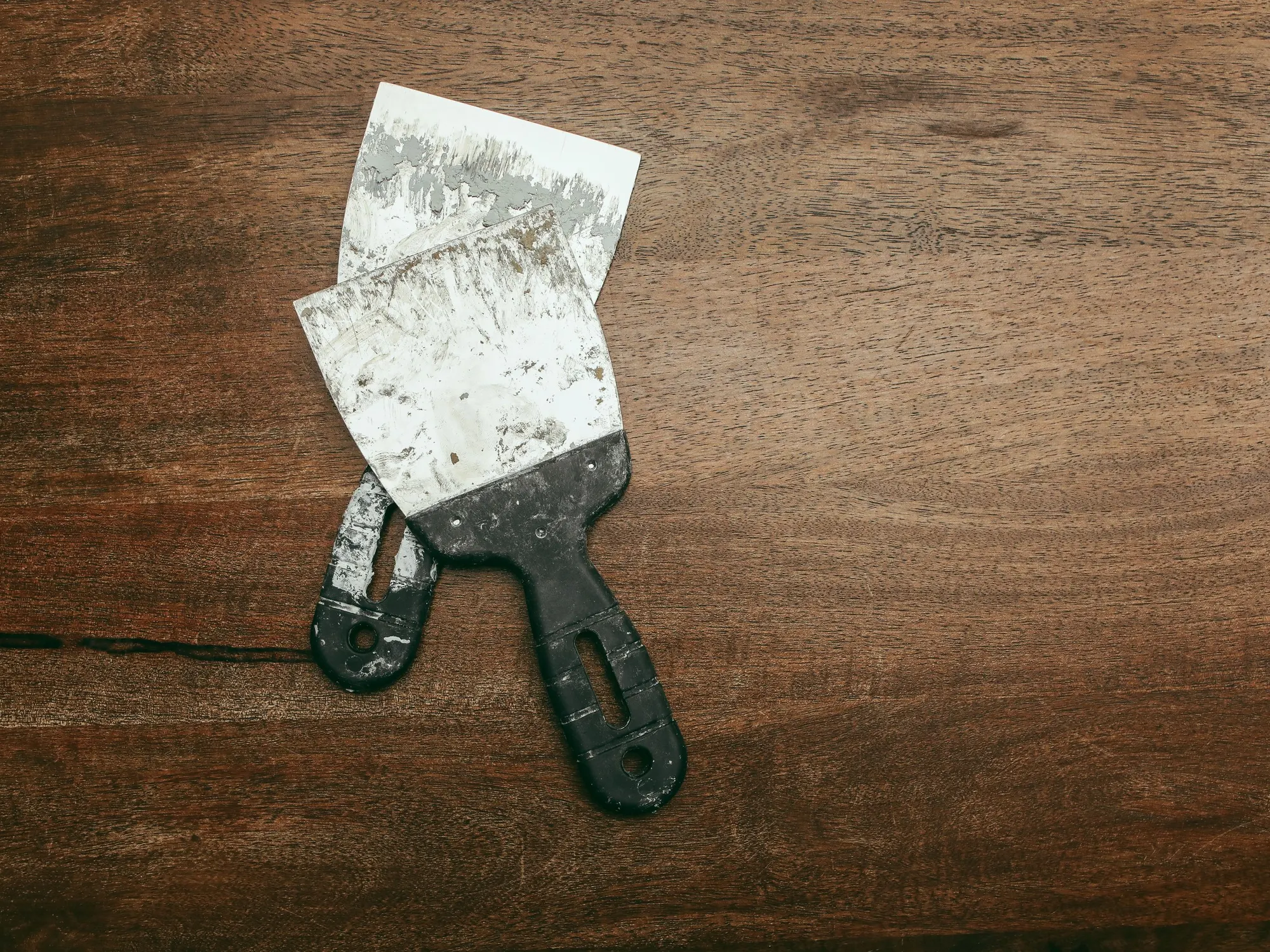 Two paint crusted putty knives lying on a dark wooden surface