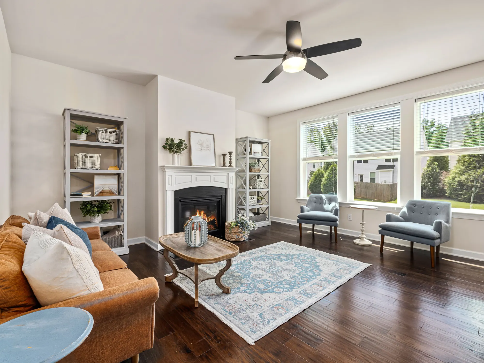 A bright living room with a white fireplace, built-in shelves, a ceiling fan and three tall windows