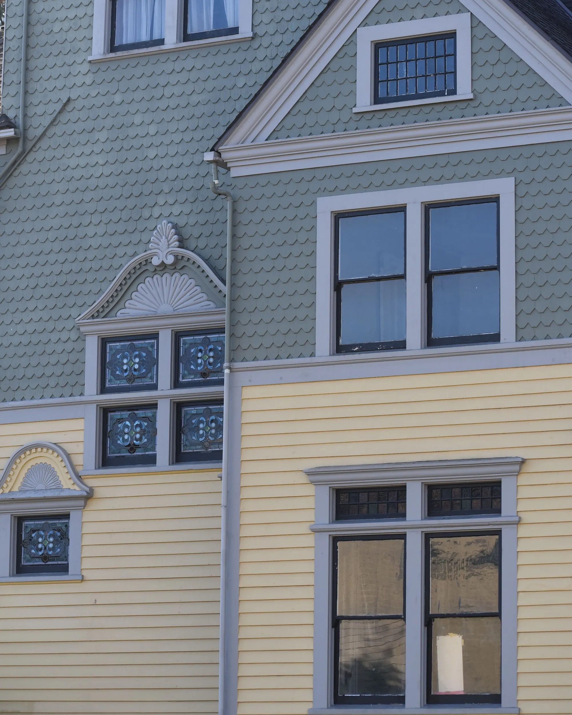 A gabled house front with grey-green fish-scale shingles above pale yellow clapboard and white-trimmed windows
