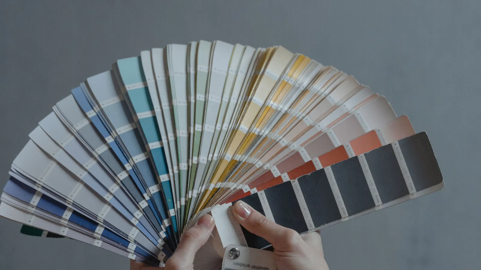 Two hands holding an open fan of paint color sample strips in blues, greens, creams, corals and grays against a gray wall