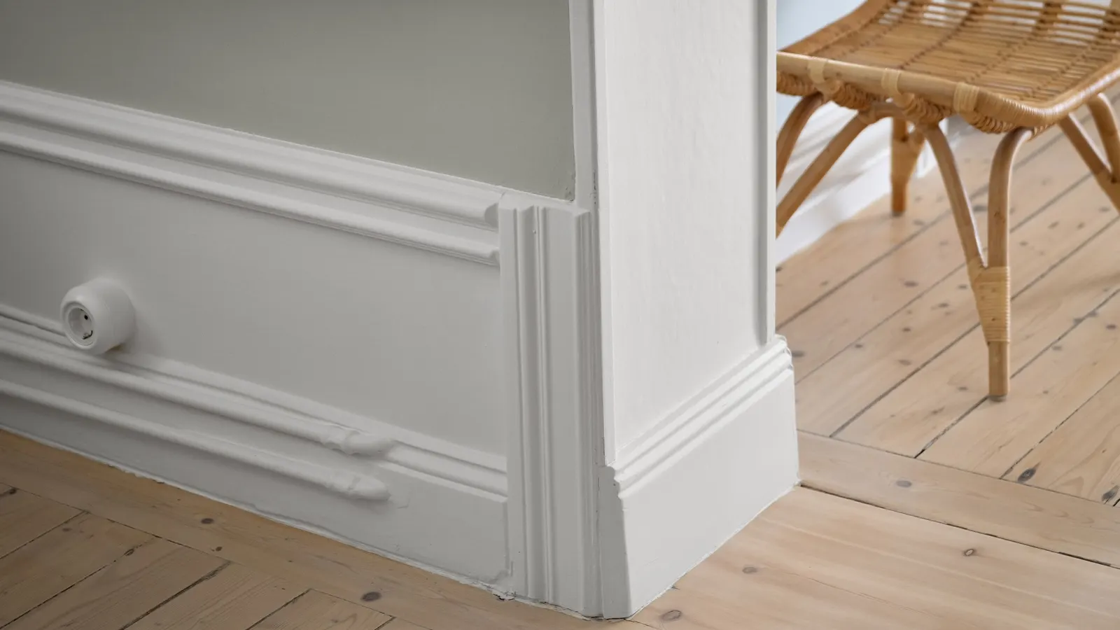 A white painted baseboard, wall moulding and door casing meeting a pale pine floor, with a rattan stool in the next room