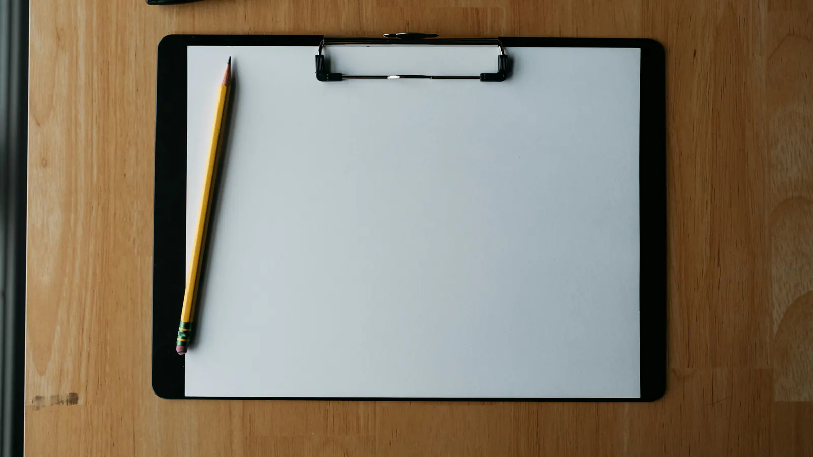 A black clipboard holding a blank white sheet with a yellow pencil, lying on a light wood desk