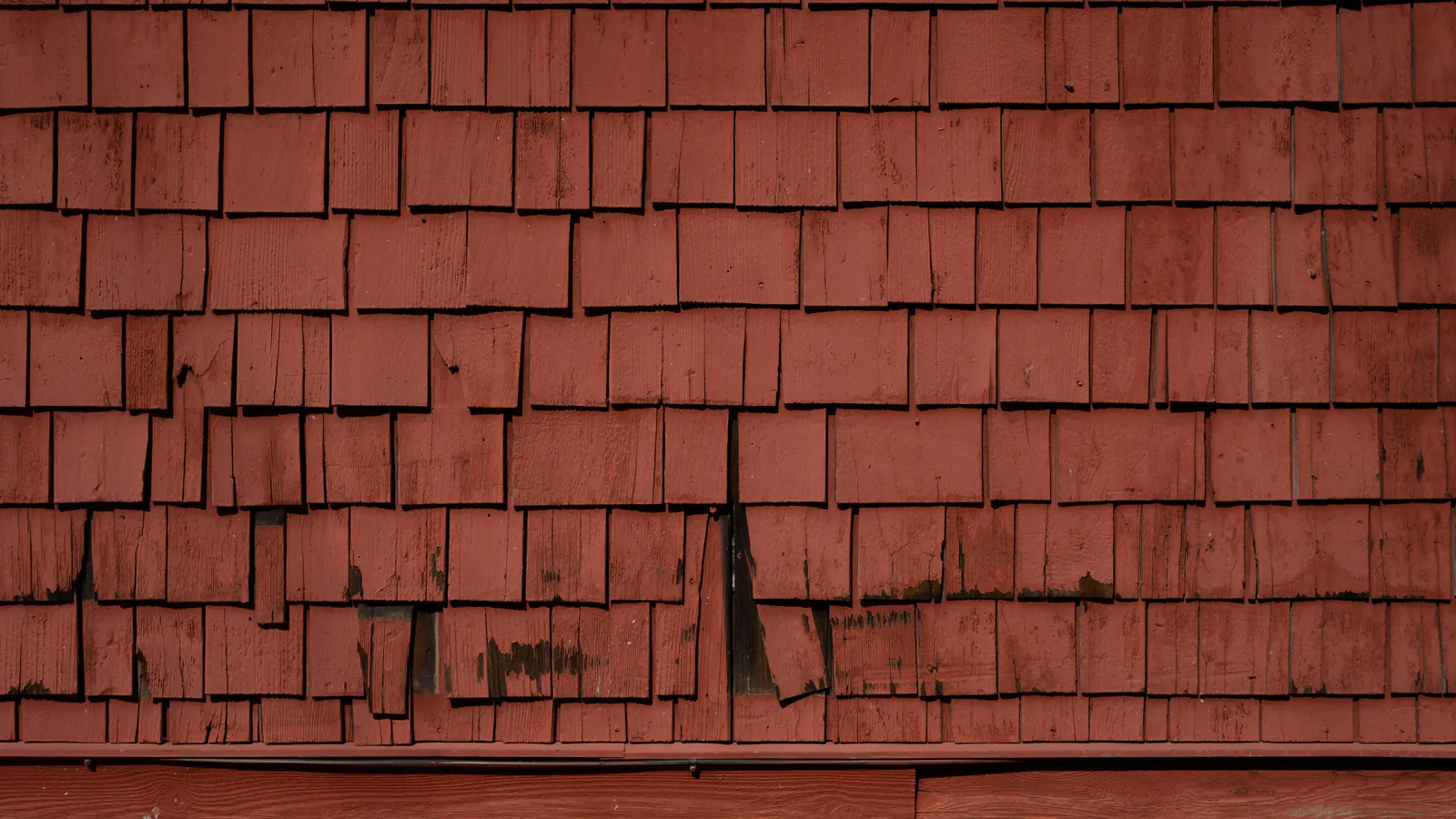 A wall of weathered red-painted wood shingles with faded, worn paint, a few split and lifting shingles and dark gaps between courses.
