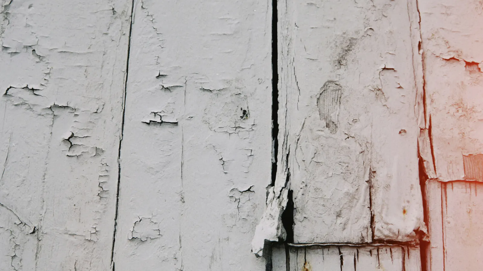 Close view of white painted wood boards with flaking, cracked paint, an open gap at the joint and a split, weathered corner piece.