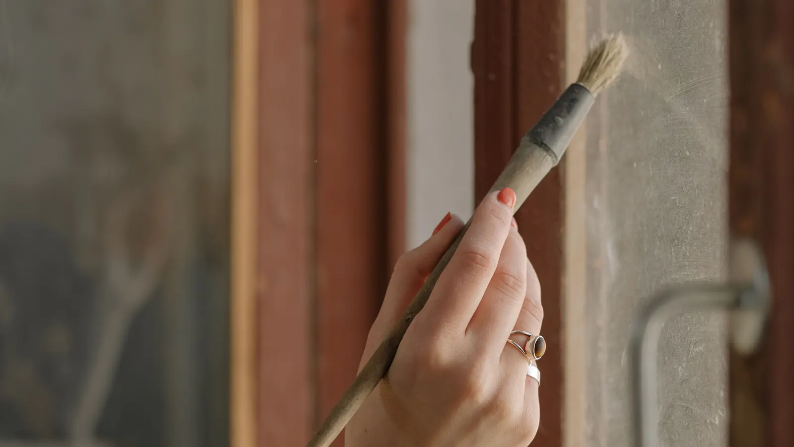 A hand holding a natural-bristle paintbrush works along a reddish-brown wooden window frame, with soft light and a blurred room behind.