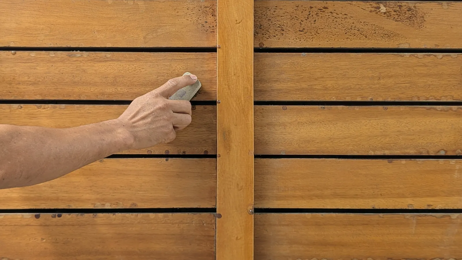 A bare forearm and hand sand a horizontal board on a warm-toned wooden slatted wall with a vertical batten crossing the boards.