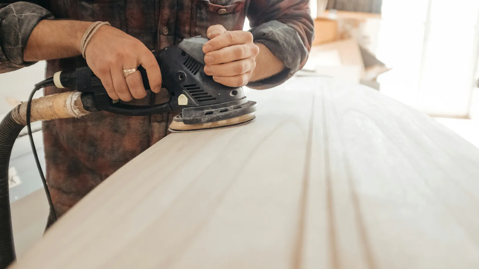 Hands guide a random orbital sander with a dust hose across a smooth, pale wood surface in soft window light.