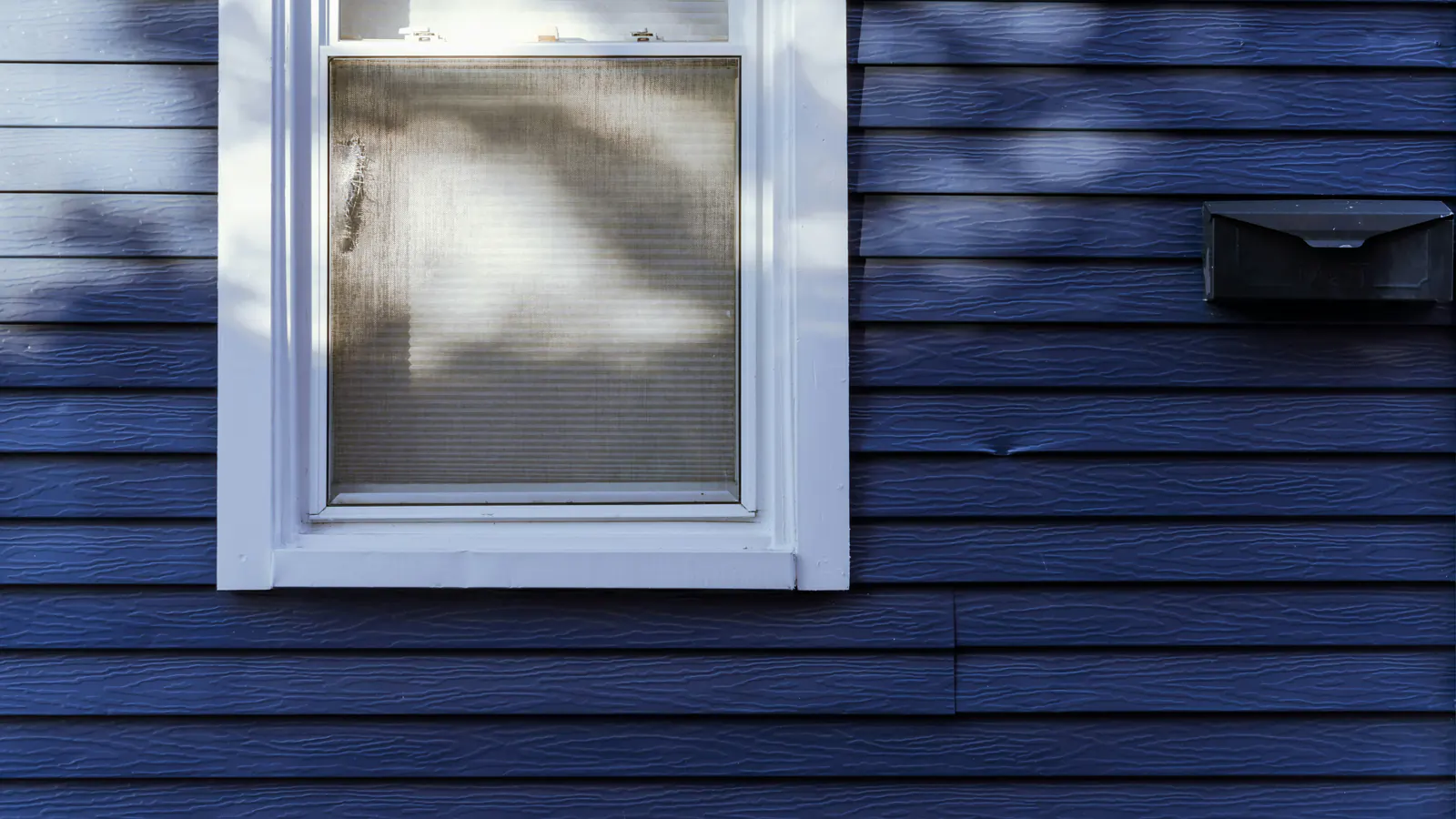White window casing set into dark blue horizontal lap siding, with dappled shade, showing the joints where trim and siding boards meet.