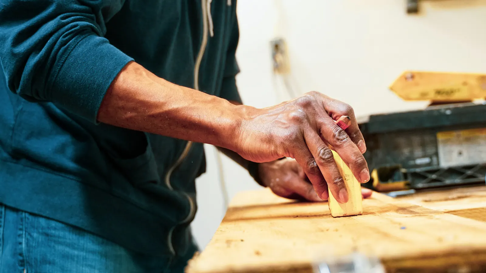A hand pressing a sanding block along the edge of a wood surface on a workbench, with tools out of focus behind.
