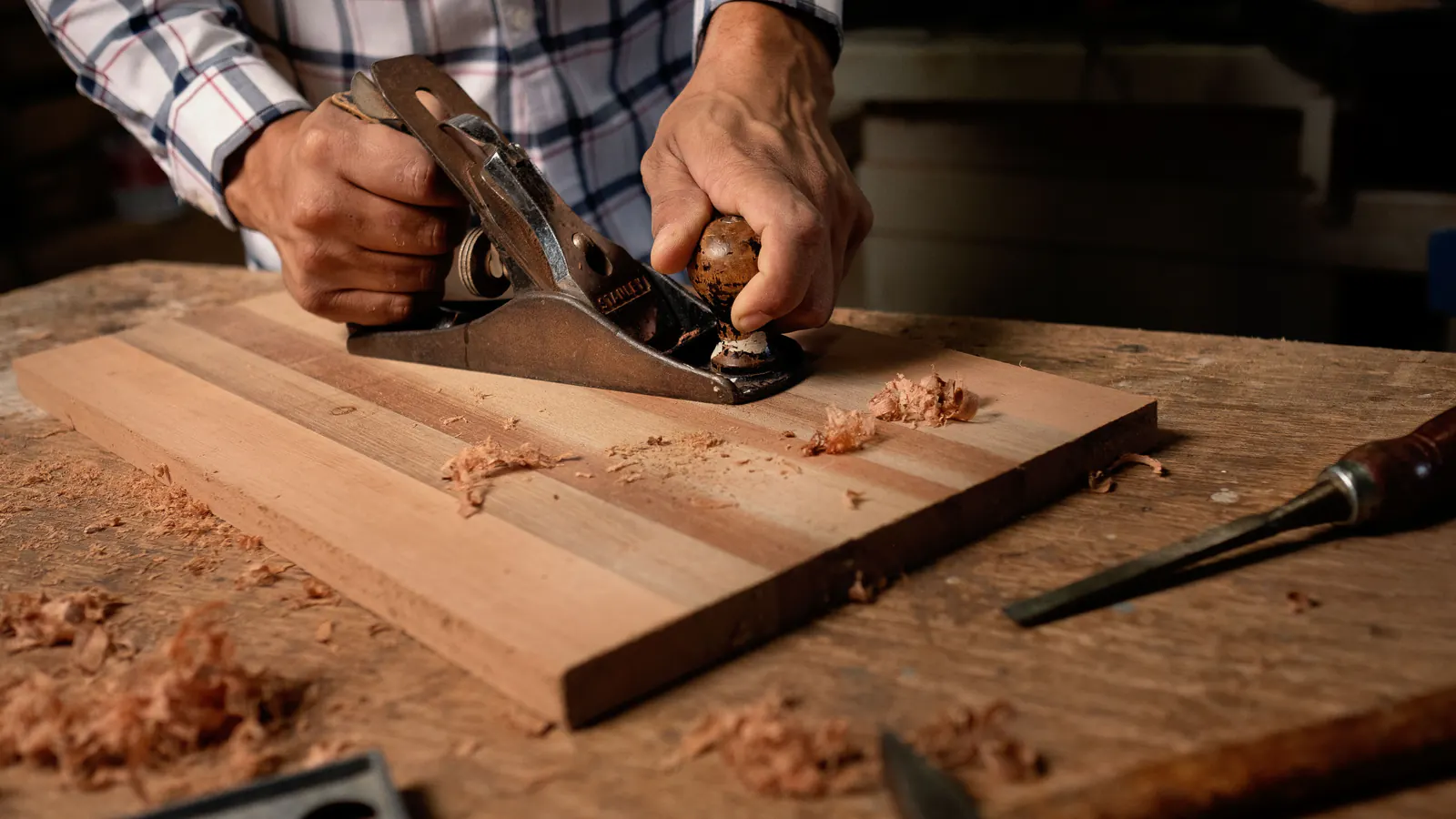Two hands pushing a hand plane along a new wood board on a workbench, with curled shavings and a chisel beside it.