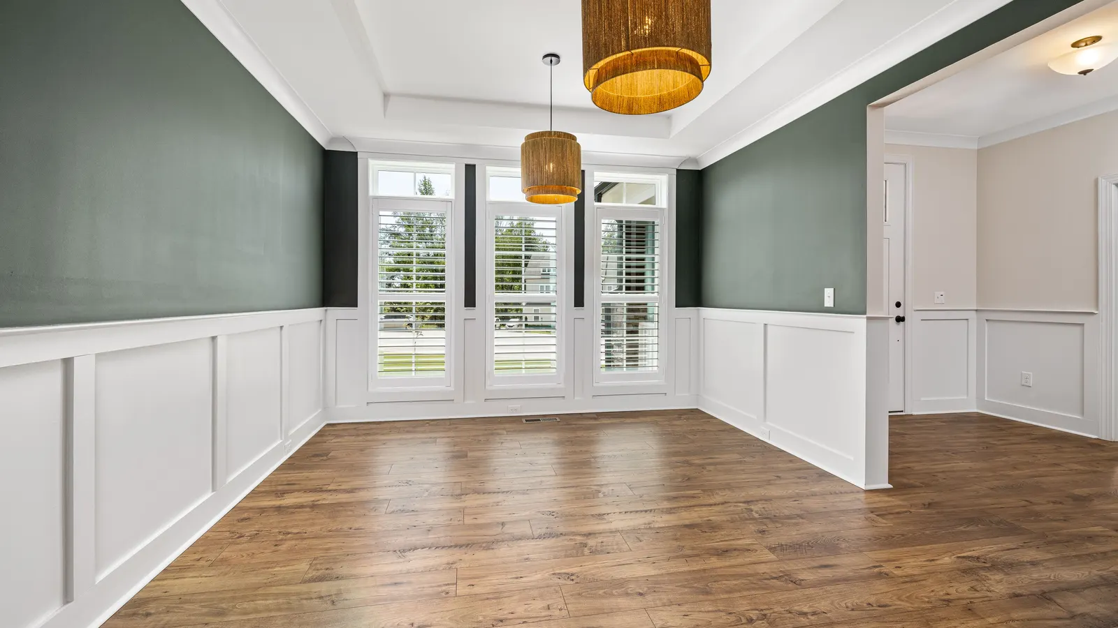 An empty dining room with sage green walls above white picture-frame wainscoting, white window trim and a tray ceiling.