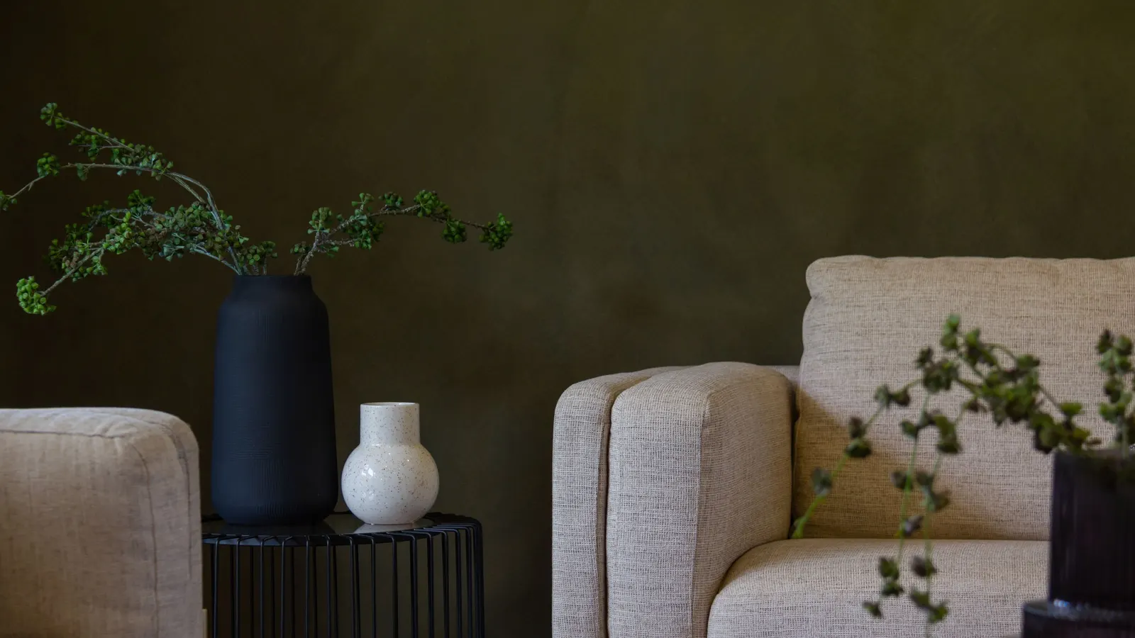 A living room corner with pale sofas, a black side table and vases in front of a mottled olive plaster wall.