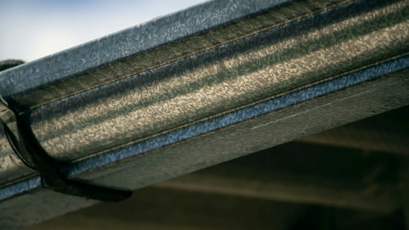 Low-angle close-up of a galvanized metal gutter running under a roof eave, held by a dark hanger strap, against a pale sky.