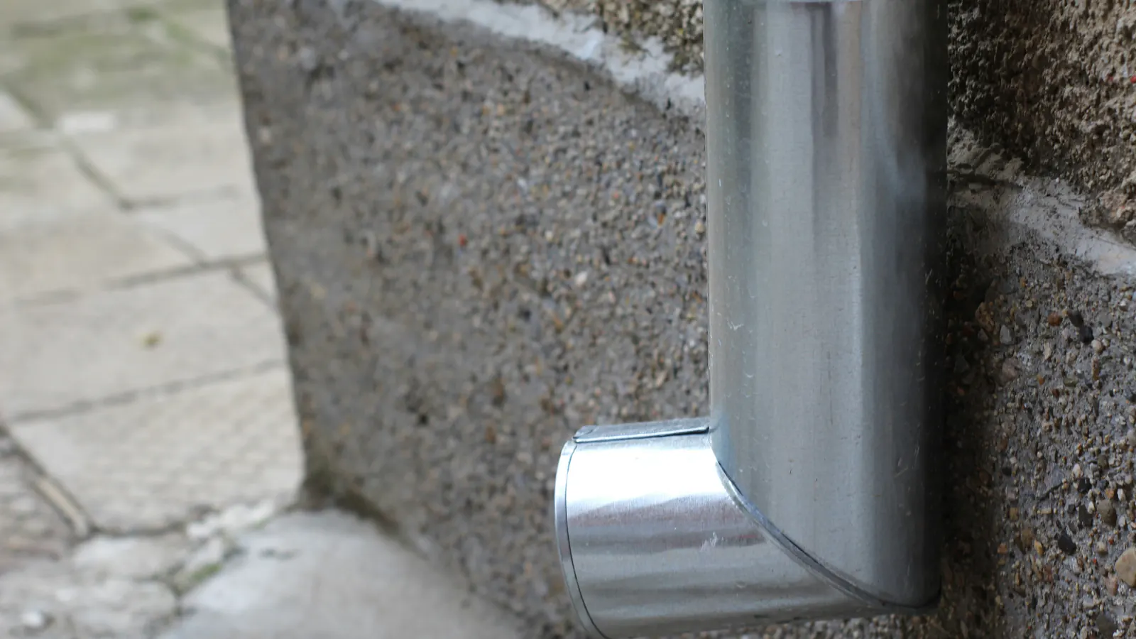 A galvanized metal downspout ending in an elbow outlet at the base of a concrete wall, just above a paved path.