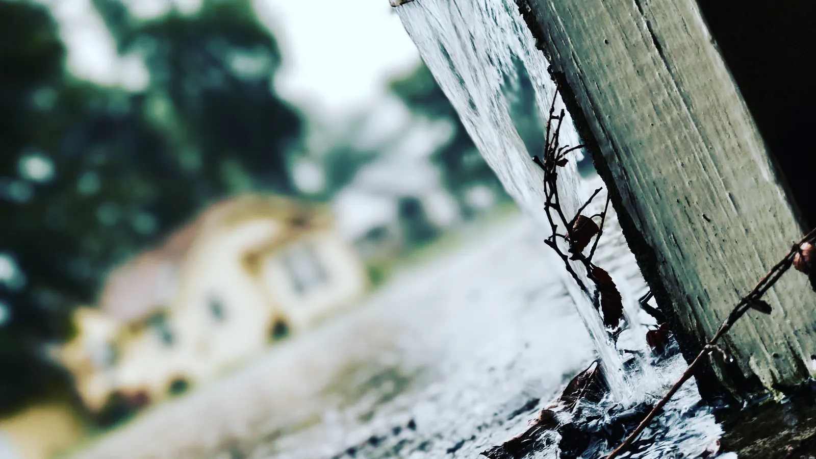 Rainwater pours in a sheet over the edge of a roof and gutter during a storm, with wire in the foreground and a house blurred behind.