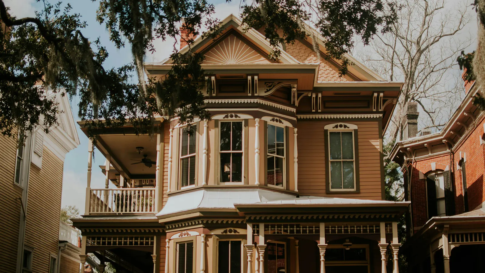 A painted Victorian house with a deep porch under oak branches