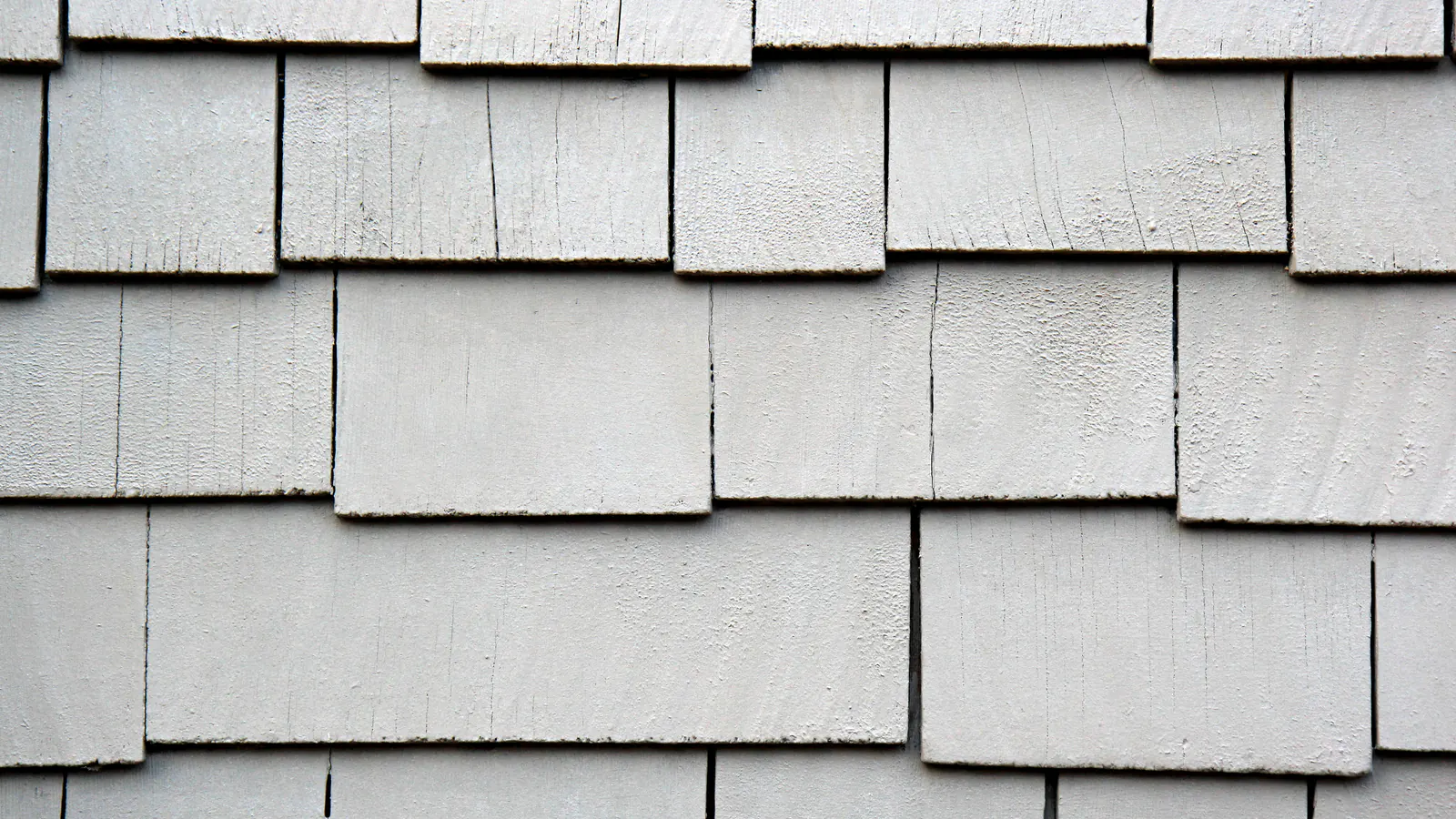 Close view of overlapping gray painted wood shingles on a wall, several with cracks and split edges