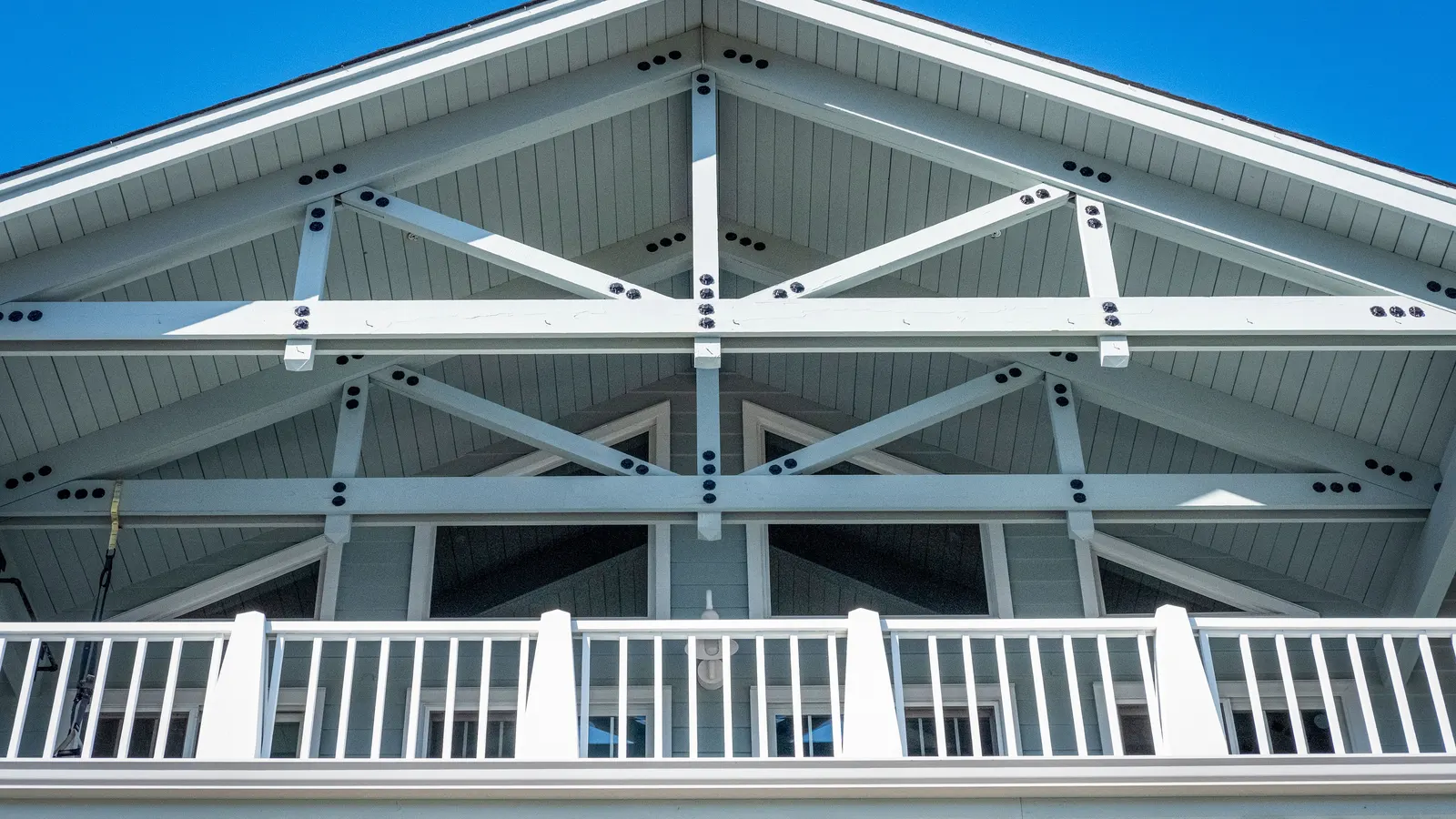 Looking up at white painted gable trusses and eave boards above a porch railing, against a blue sky