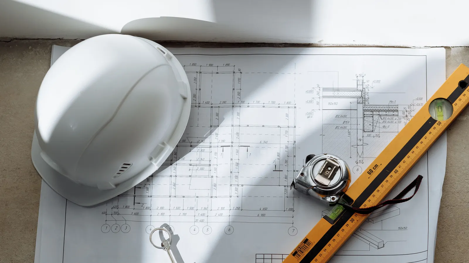 A white hard hat, a key, a tape measure and a spirit level on a printed house plan in sunlight