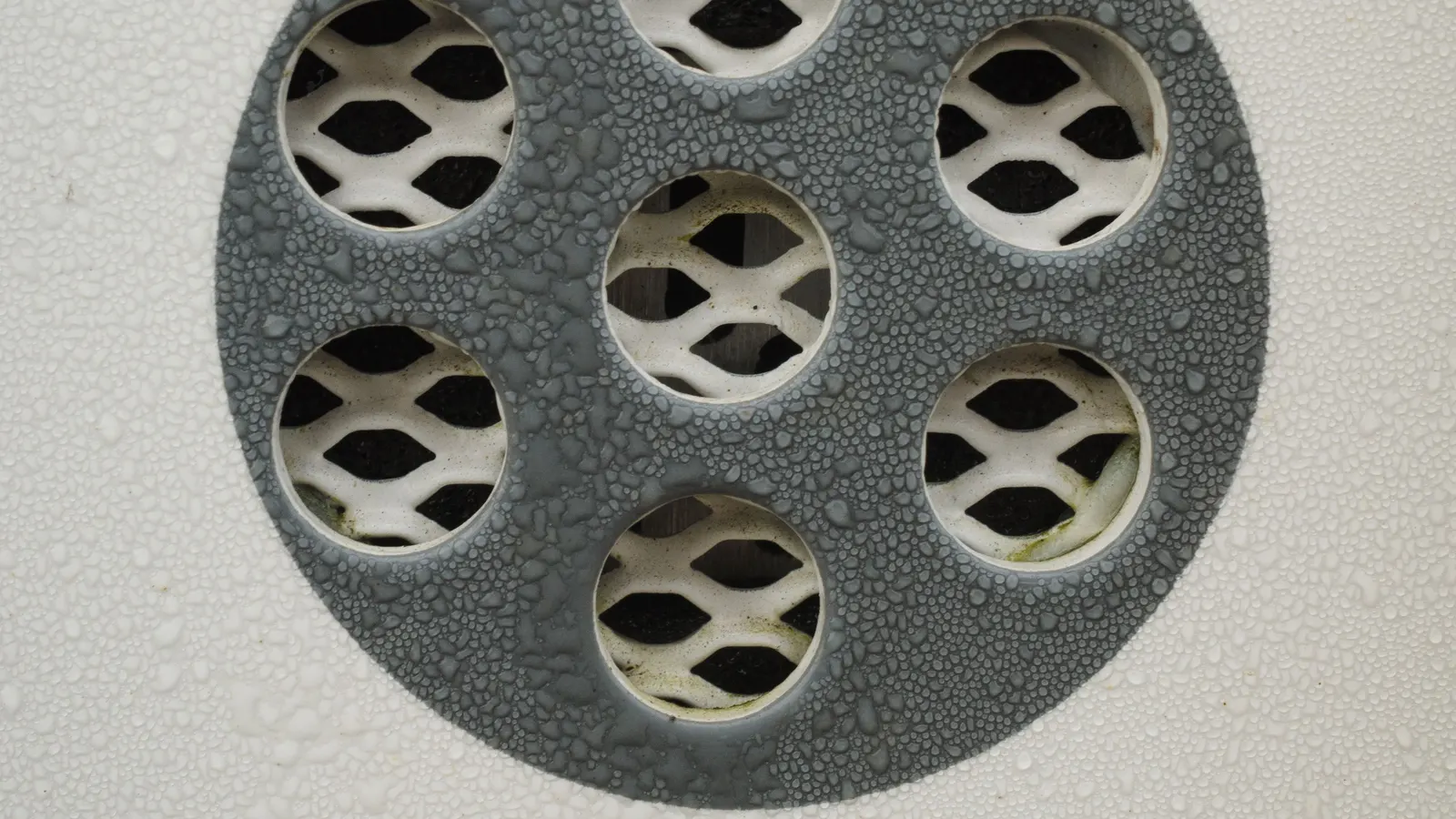 A round gray cover with mesh openings set in a pale wall, beaded with water