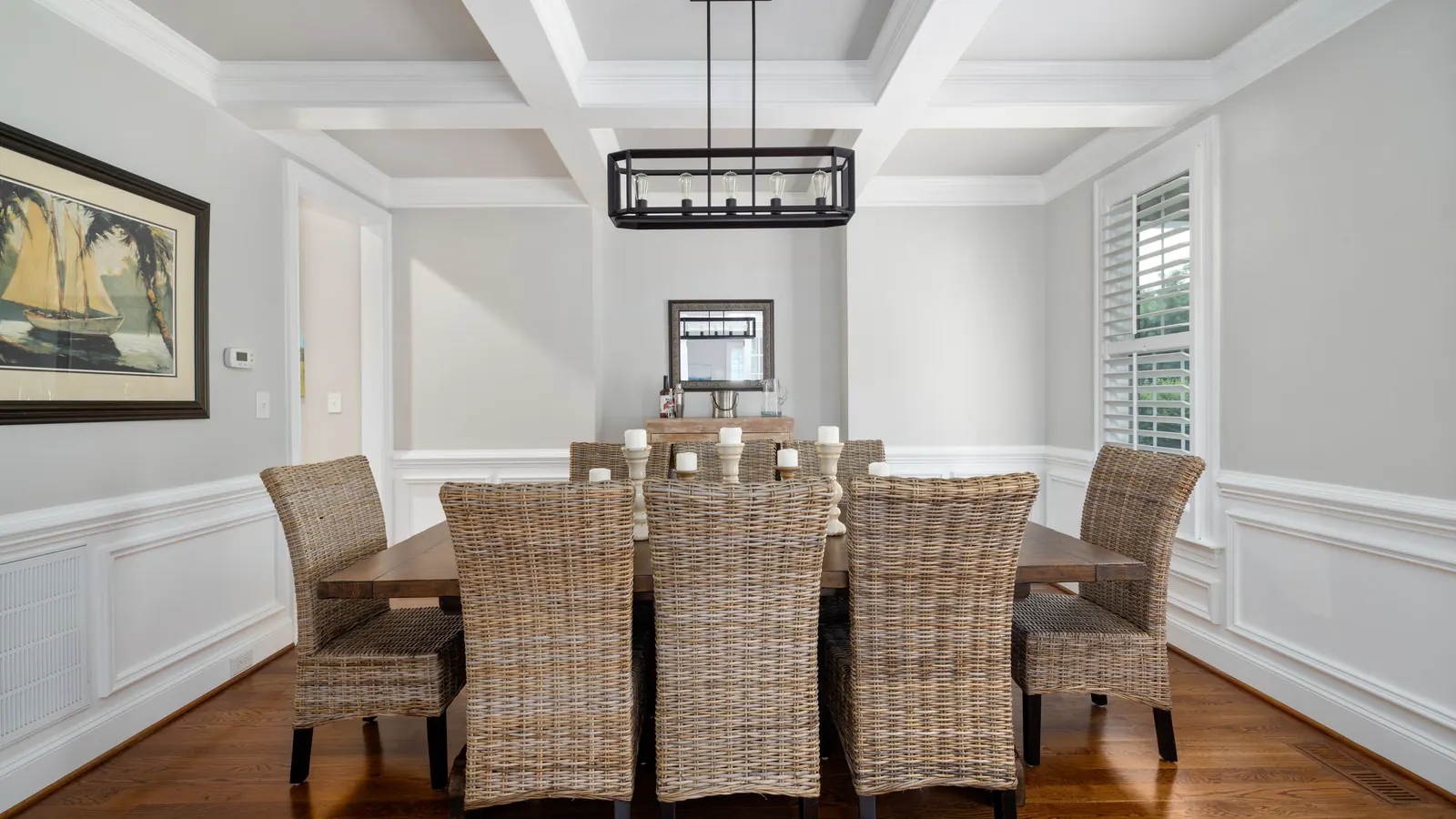 A dining room with a white coffered ceiling, wainscot paneling, crown moulding and plantation shutters, set with a wood table and wicker chairs
