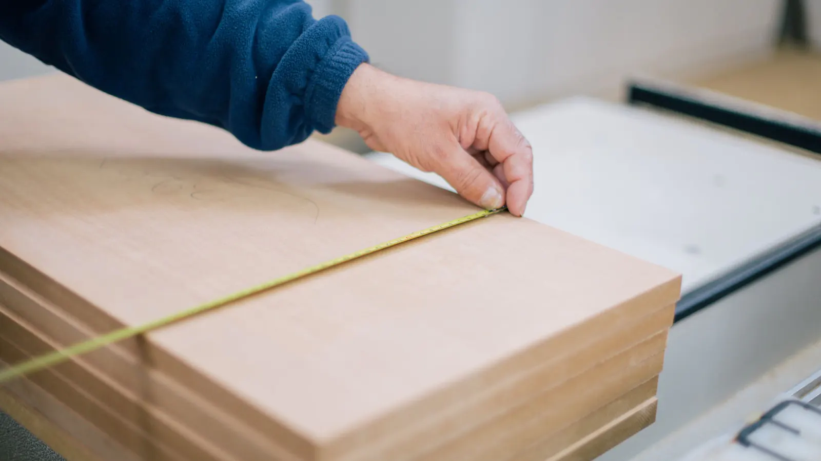 A hand stretching a tape measure across a stack of pale wood panels in a workshop