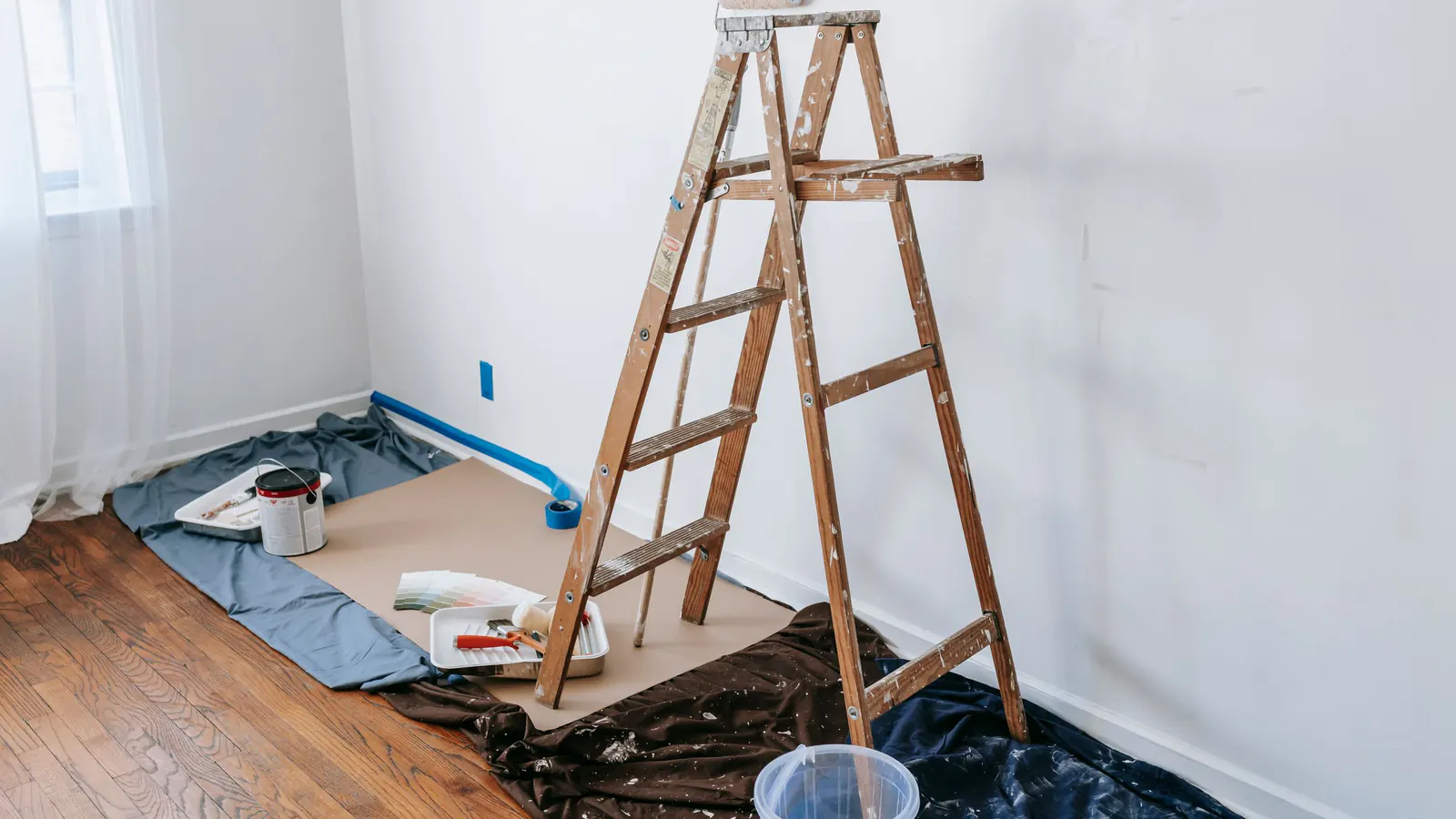 A wooden stepladder in the corner of a white room, standing on drop cloths and paper floor protection, with blue tape along the baseboard and brushes on a tray