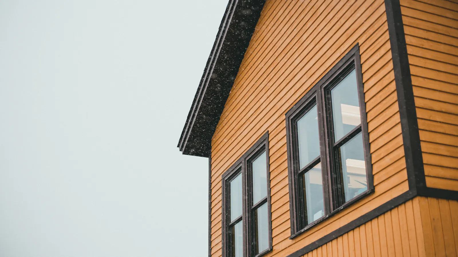 Ochre lap siding with dark brown window casings and corner trim on a gable wall, under a pale winter sky