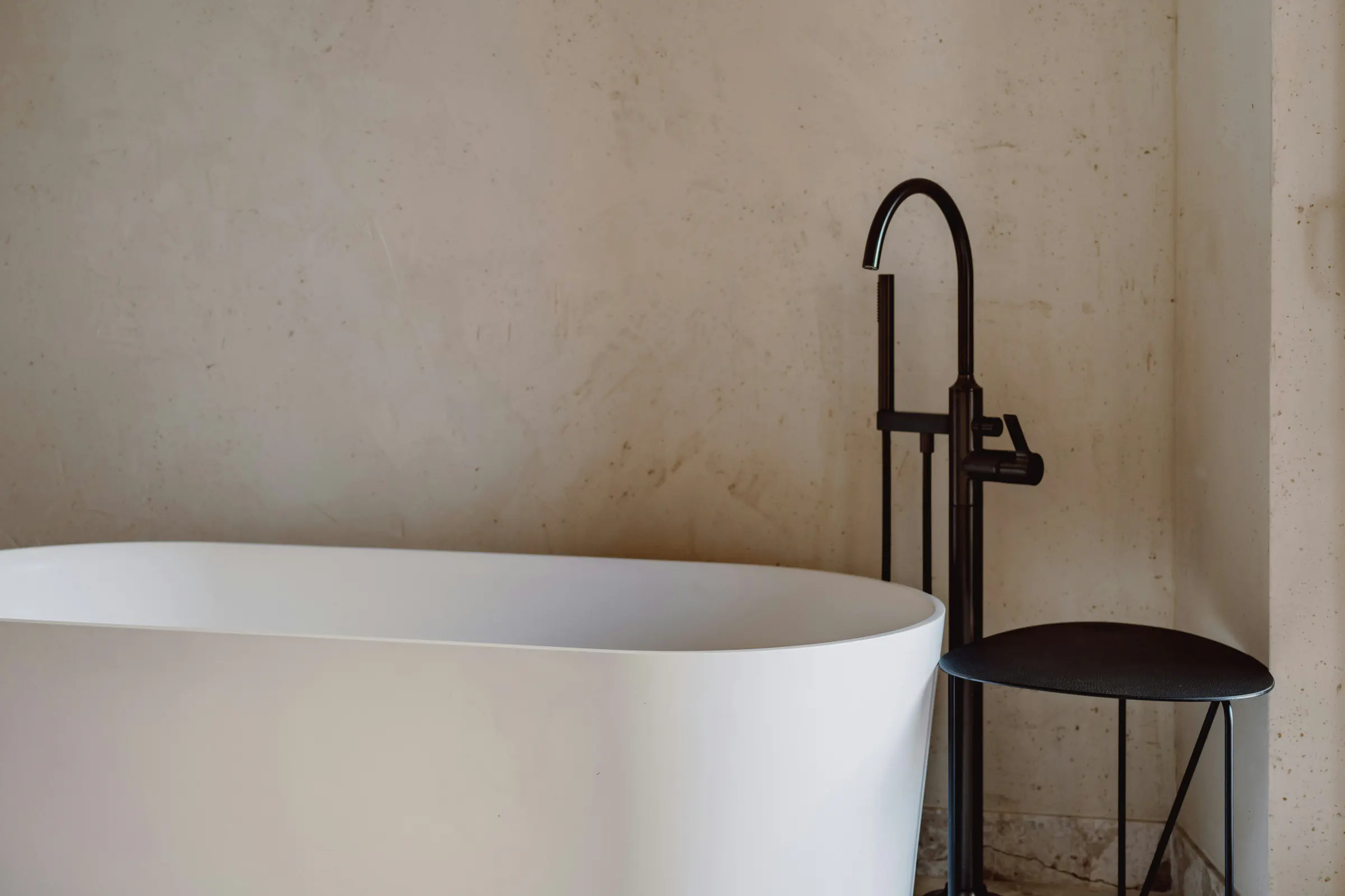 A white freestanding tub and a black floor tap against a limewashed plaster wall over a terrazzo floor