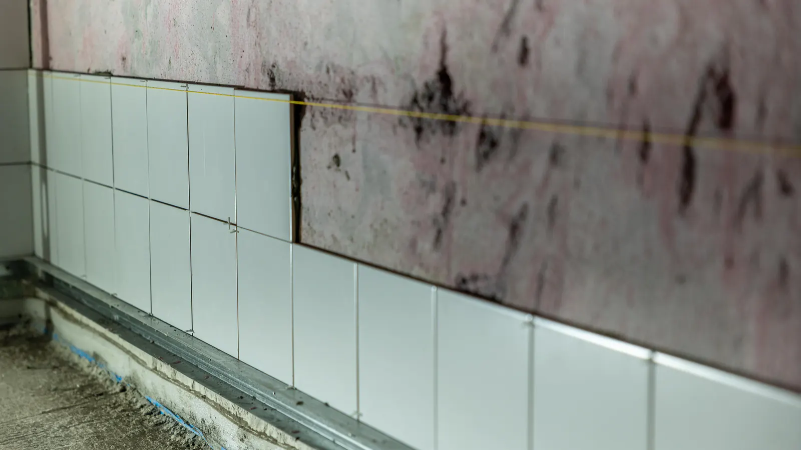 White wall tiles set partway up a bare wall along a taut yellow level line, above a rough concrete floor