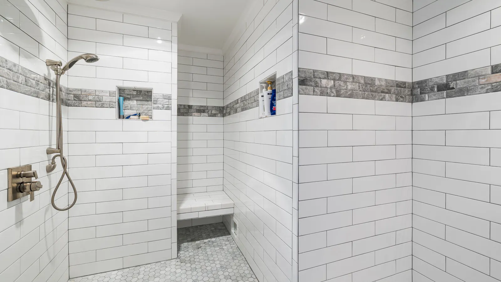 A walk-in shower in white subway tile with a grey stone band, a tiled bench, a wall niche and a hexagon mosaic floor