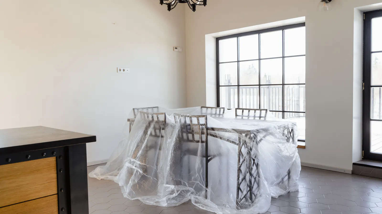A bright empty dining room with the table and chairs fully wrapped in clear plastic sheeting under a black chandelier beside tall windows