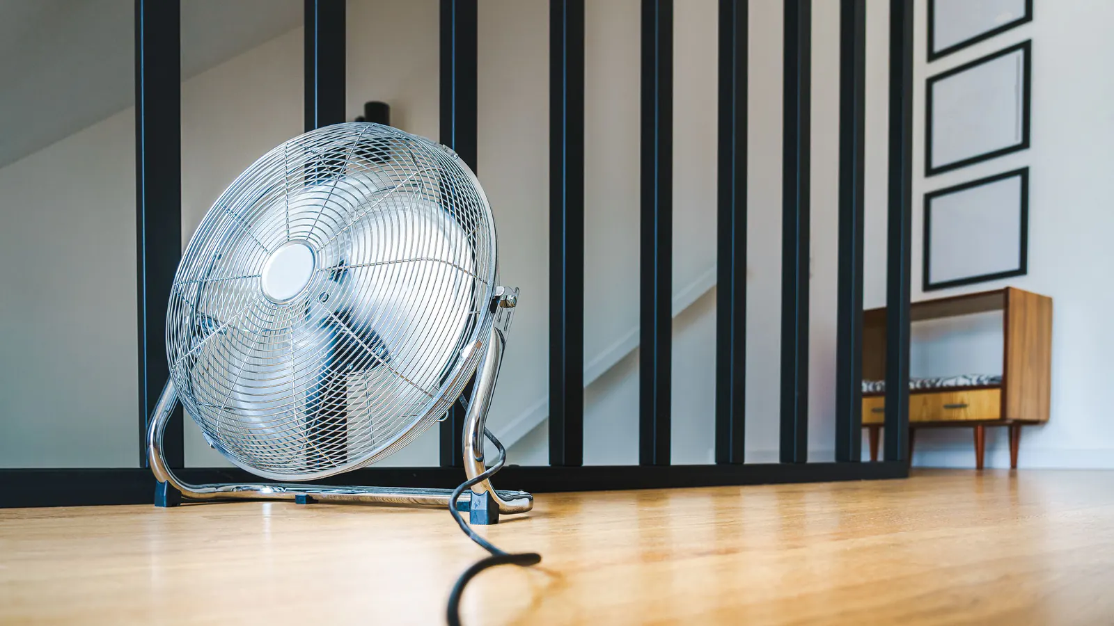 A chrome high-velocity floor fan running on a warm wood floor in a bright room with black stair balusters behind it