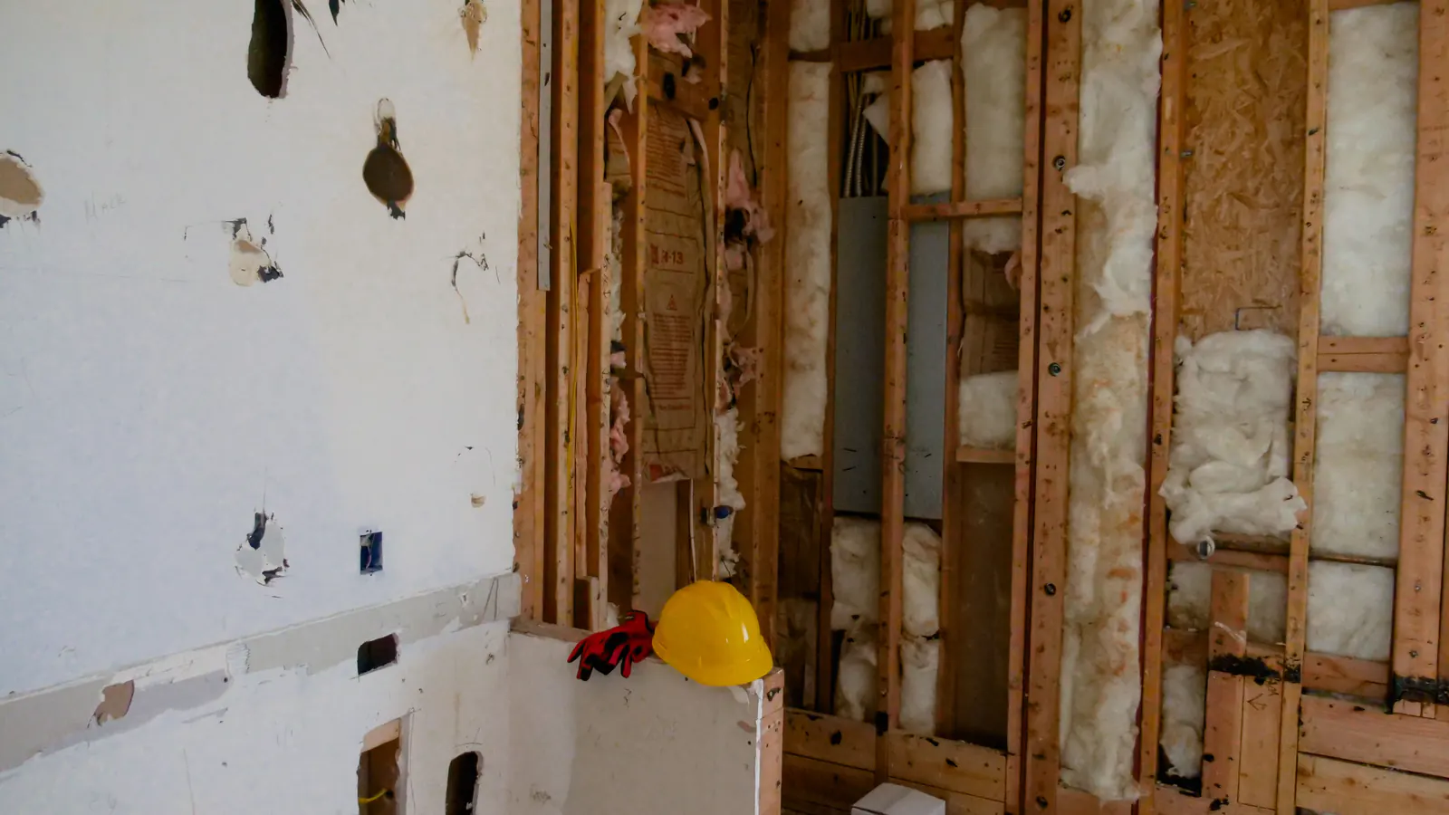 A room with the drywall partly torn away, exposing wood studs, insulation and old wiring, with a yellow hard hat resting on a ledge