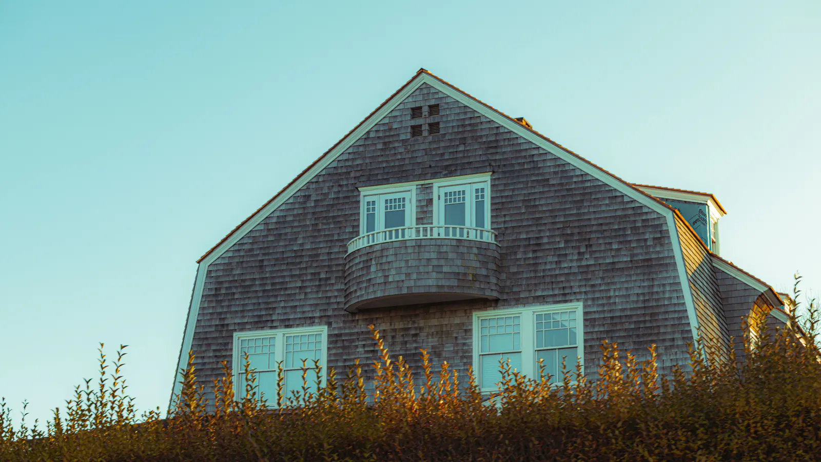 A cedar shingle house with a tall gable and a curved balcony against a blue sky