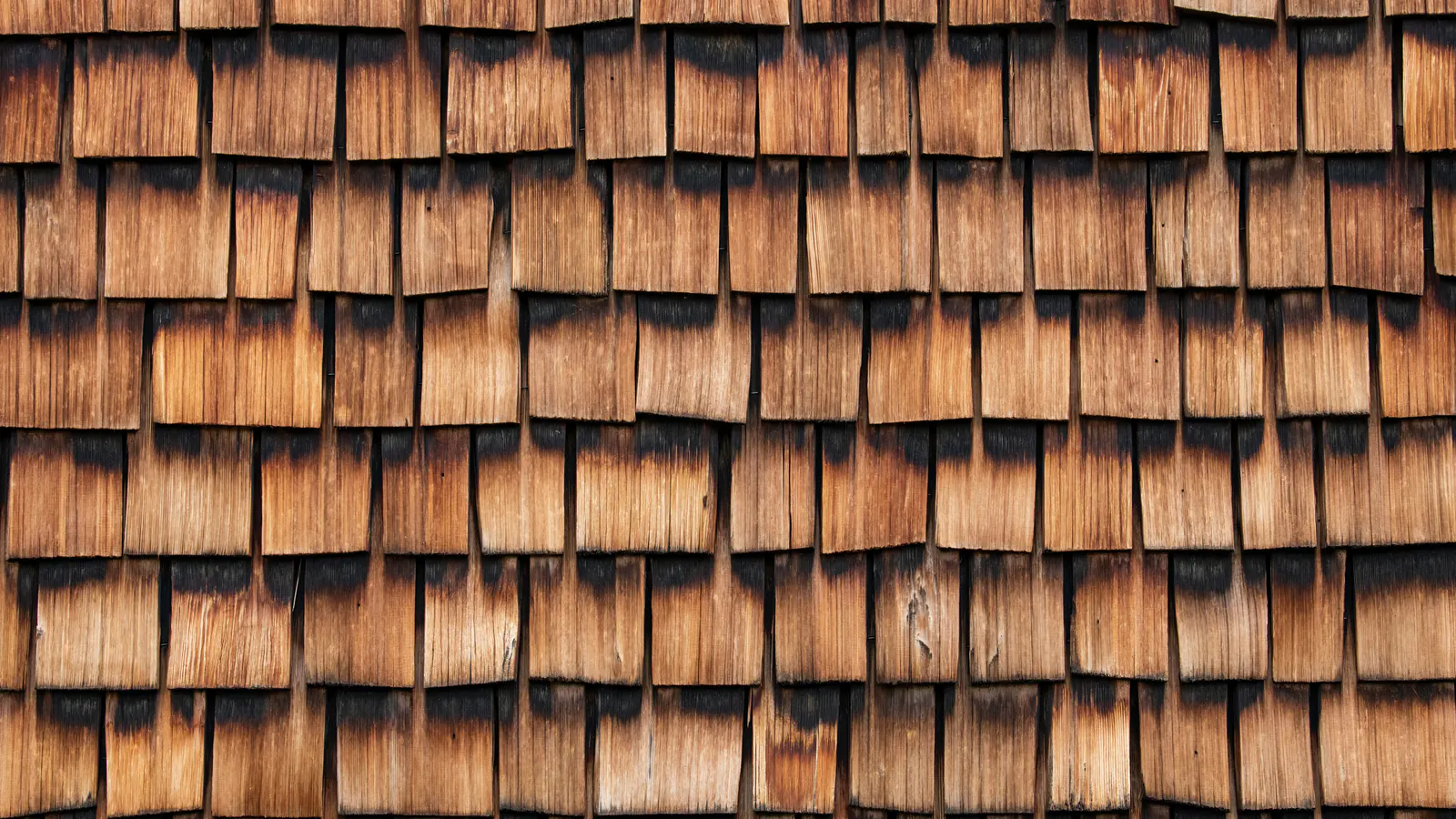 A wall of warm cedar shingles with lighter faces and darker weathered lower edges, showing uneven, sun-worn color from course to course