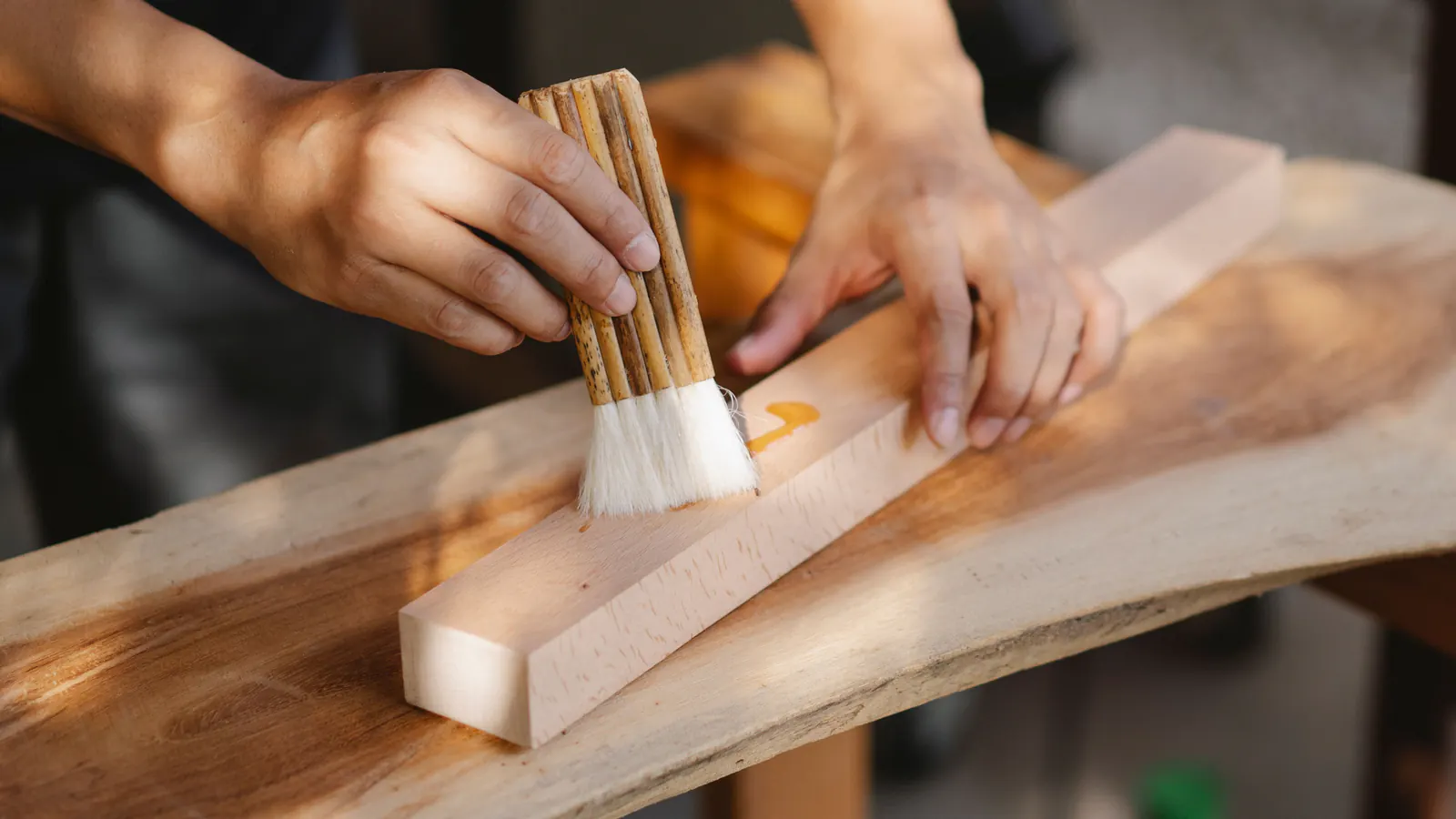 Hands holding a wide natural-bristle brush and working a finish into the grain of a pale wooden plank on a wood slab